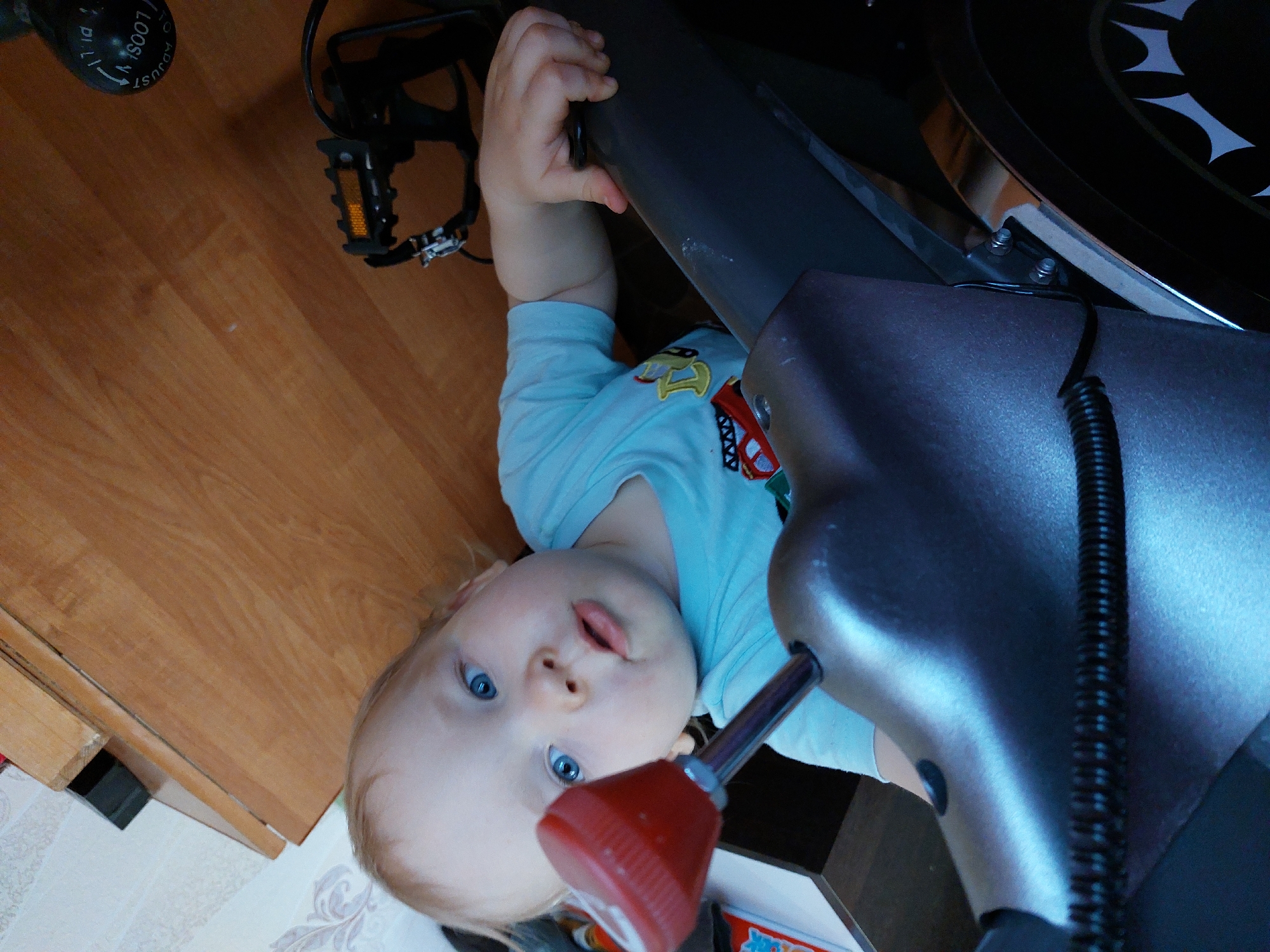 A low-angle shot depicts a baby in a light blue shirt with cartoon patches, lying underneath an exercise bike. The baby has blonde hair and blue eyes and is looking directly into the camera. The baby's left hand is holding onto a gray metal part of the exercise bike. The bike's pedal is visible on the left, and part of the bike's flywheel can be seen on the upper right. The floor is a combination of wood planks and patterned tile. The setting is indoors. There is no indication of the location being Большой Букор, Russia.