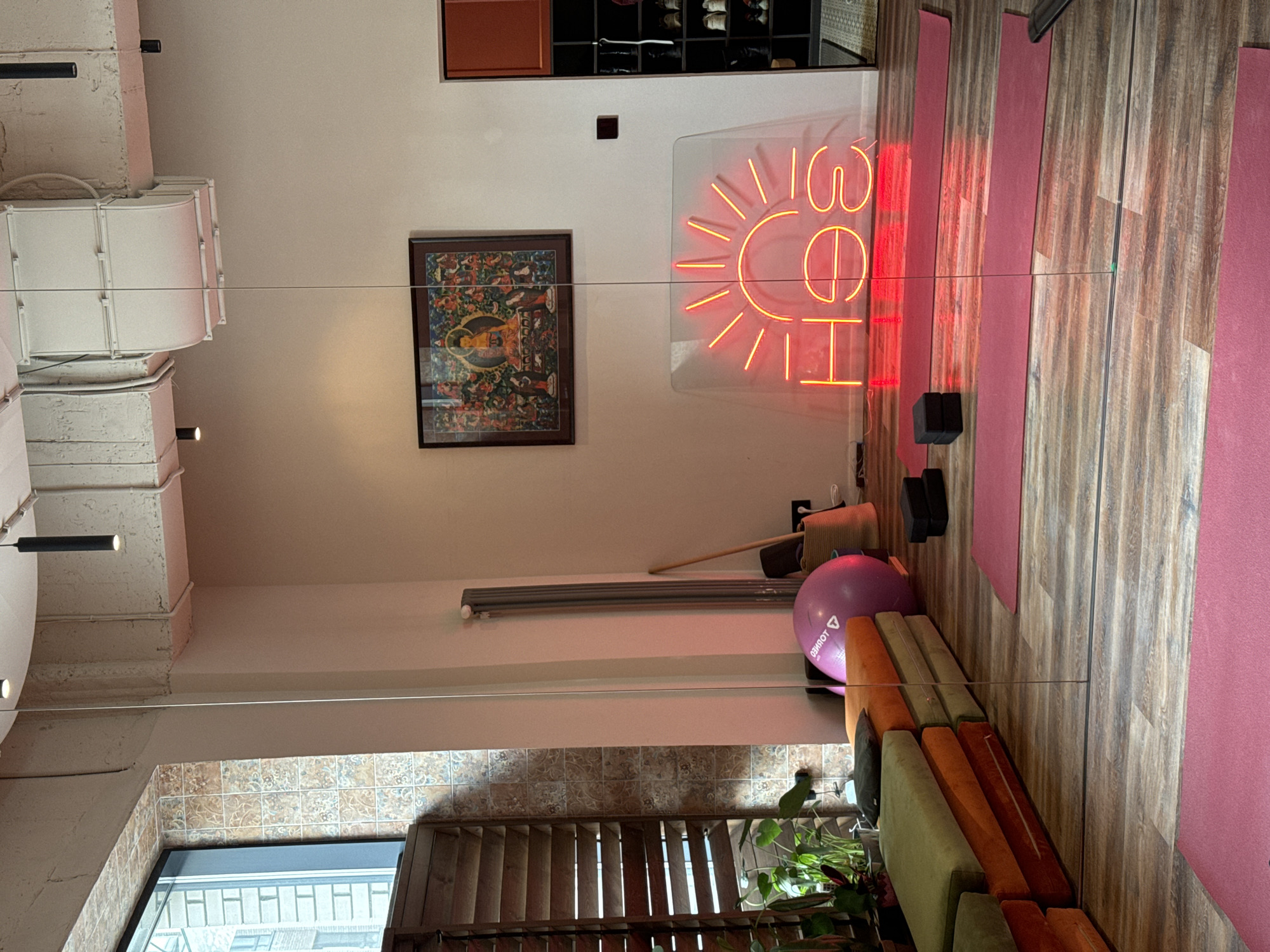 This indoor shot features a yoga studio. On the left, an open window reveals an urban setting, possibly in Krasnoyarsk, Russia, though there are no overt visual cues confirming the city. The wall adjacent to the window is tiled and covered with horizontal blinds. A row of low seating, with sections in olive, orange and brown, is set against the wall, near a plant. Above, on the wall, there are stacked yoga bolsters, a balance ball, and a storage rack. Moving to the right, the room features decorative elements including a framed art piece and a brightly lit neon sign. Dark yoga blocks are stacked up next to the floor, on yoga mats. The floor features individual yoga mats. A ventilation system runs along the ceiling.
