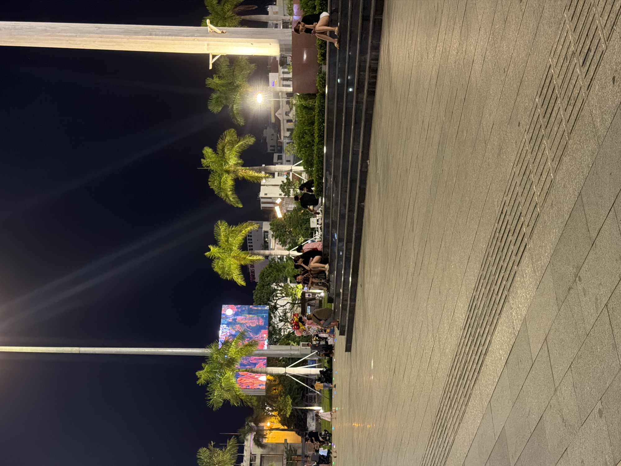 The image depicts a nighttime scene at an outdoor plaza in Da Nang, Vietnam. Several people are visible, some sitting on broad, tiered steps in the foreground, while others are scattered across the paved area. A large digital billboard displaying colorful imagery is prominent in the background, surrounded by palm trees and urban architecture. Streetlights and architectural lighting illuminate the scene. The ground in the foreground is paved with large, rectangular tiles and features a textured, grooved section.