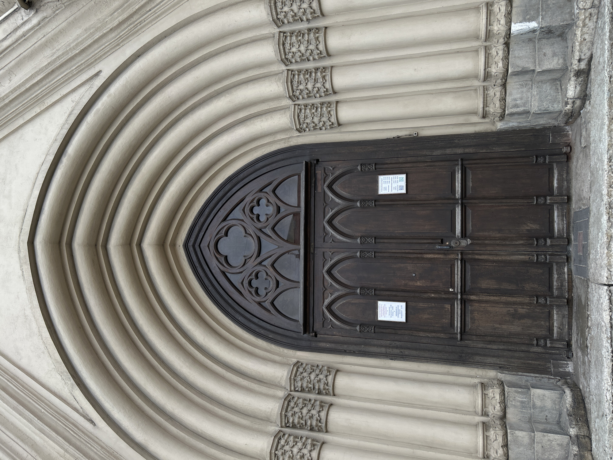 A photograph captures a gothic-style arched doorway. The archway is formed by a series of concentric, cream-colored stone arches, adorned with decorative carvings. The double doors are made of dark wood and feature a pointed arch design with quatrefoil detailing. Two small white signs are affixed to the doors. The doorway is located in Saint Petersburg, Russia.