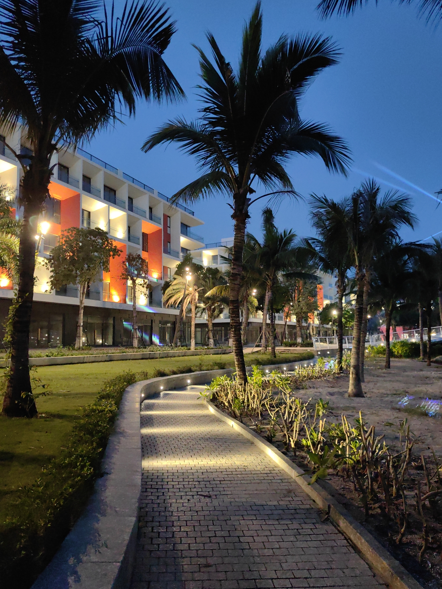 The image shows a lighted pathway in Bai Chay Ward, Vietnam. The path is made of grey bricks and curves slightly, with inset lights illuminating the way. It runs alongside a manicured green lawn on one side and an area with shrubs and trees on the other. Palm trees are prominent throughout the scene, silhouetted against a dark blue sky. In the background, a multi-story building with white walls and orange accents is visible, with lights on in some of the rooms. The overall lighting suggests it is dusk or night.