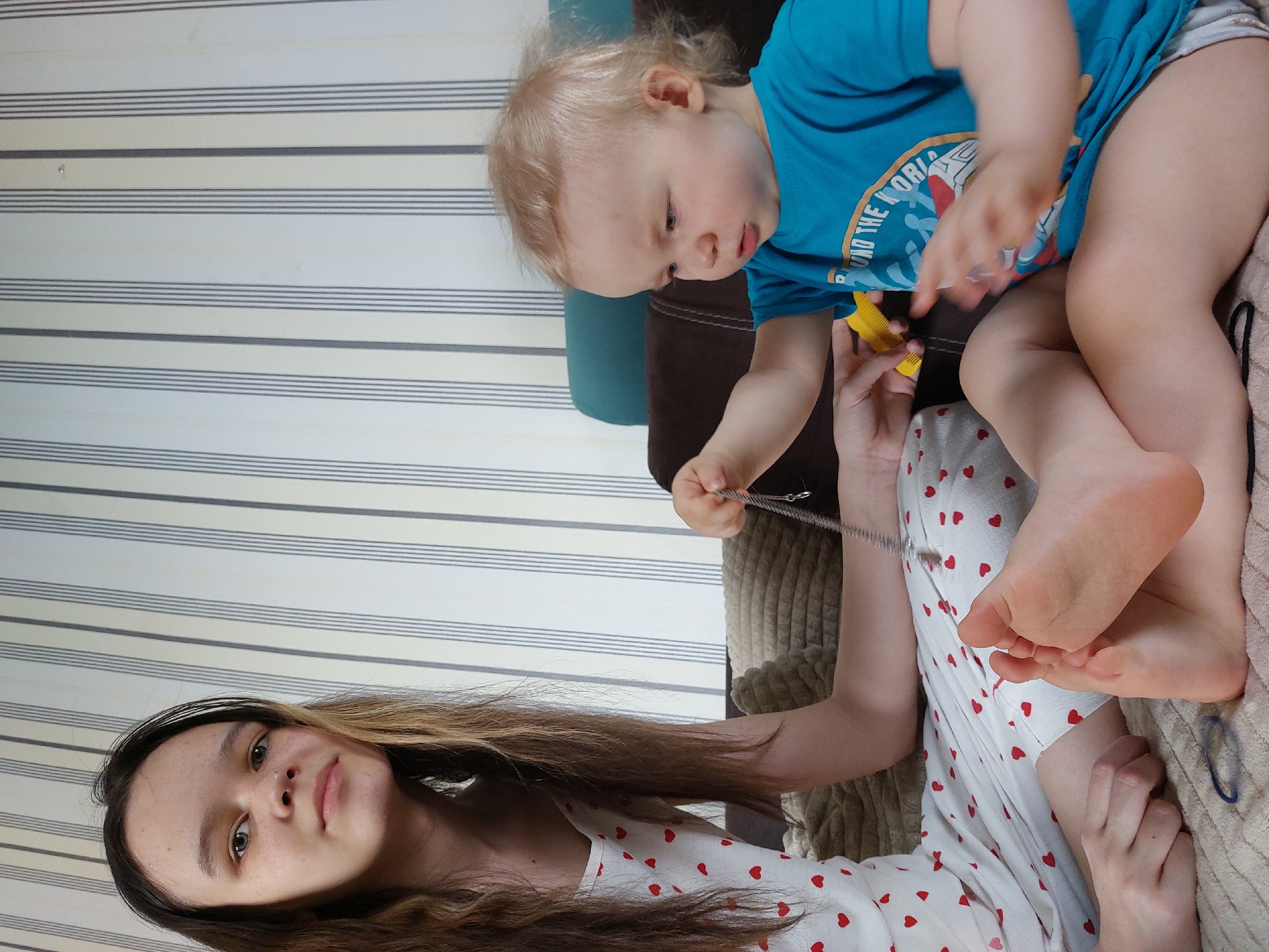 The image shows a young woman and a toddler in an indoor setting. The woman is lying down, wearing a white dress with red hearts. Her long brown hair is partially visible. She is holding a silver chain necklace. The toddler, wearing a blue t-shirt and diaper, is sitting on the woman's lap and playing with a yellow comb. The background features a wall with horizontal stripes. The location is Chaikovsky, Russia.