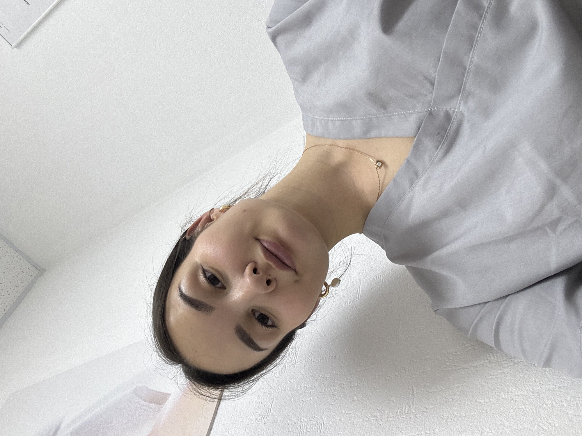 A woman is photographed from a low angle in a room with white walls. She has dark hair, fair skin, and is wearing a light gray garment that appears to be a medical uniform. She has a gold necklace and earrings. The location is possibly Chaikovsky, Russia, but there is no visual evidence to confirm this.