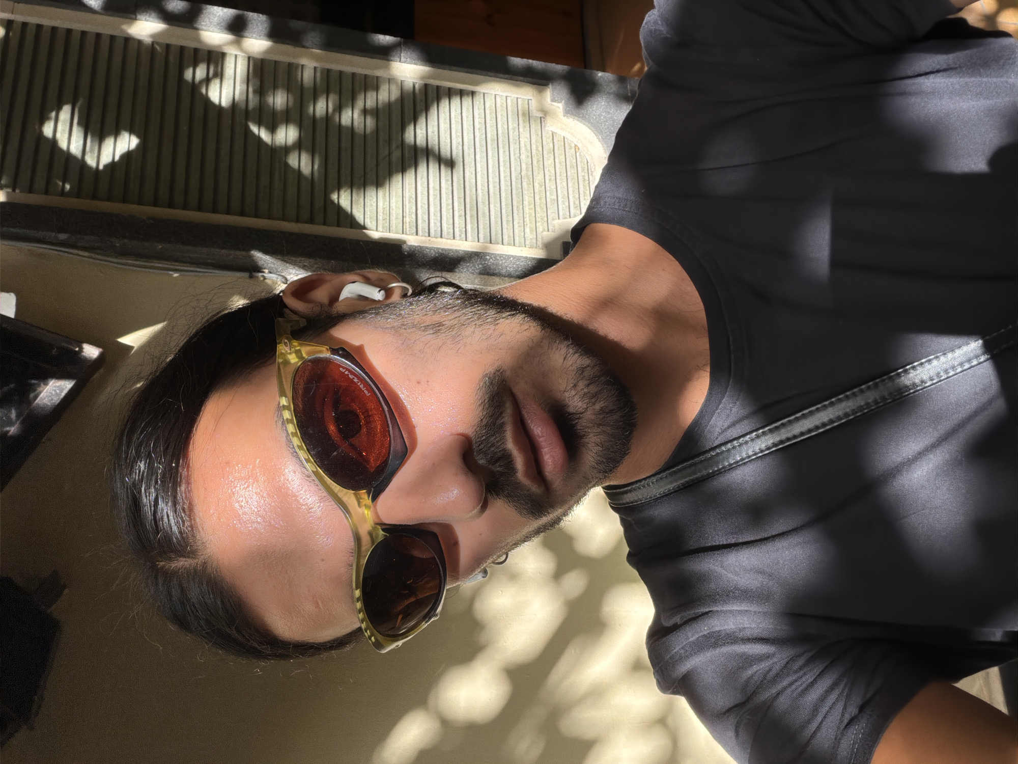 A medium close-up captures a man wearing gold-framed sunglasses with reddish-brown lenses. An earbud is visible in his ear. He is wearing a dark shirt. The background is a blur of light and shadow, with what appears to be a stone structure and the dappled shadows of leaves or branches. It is not possible to confirm the specific location in North Kuta, Indonesia from the image.