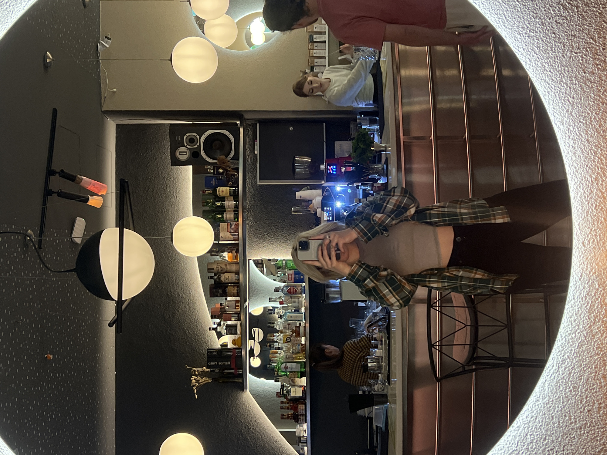 A selfie of a person in a plaid shirt taken in a bar. Behind the person is a bar with a variety of alcoholic beverages displayed on shelves. Another person is standing behind the bar, and a third person is partially visible in the background. The bar has a modern design with spherical lighting fixtures and appears to be in an upscale location. The image is taken through a mirror with a textured border. There is no evidence to suggest that the bar is specifically located in the city of Rafailovići, Montenegro.
