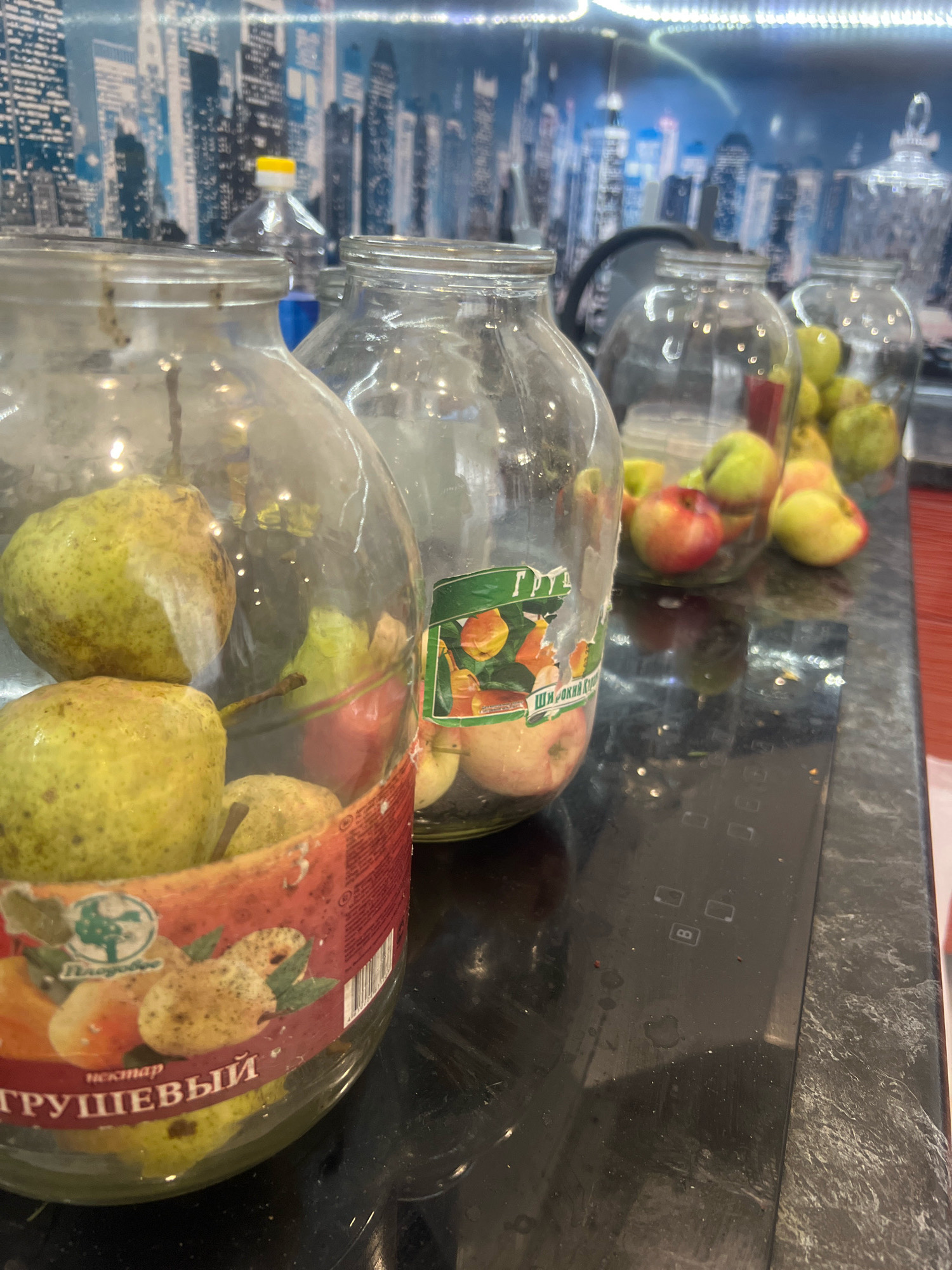 A close-up, eye-level shot displays several large glass jars filled with pears and apples, situated on a dark countertop in what appears to be a kitchen. The jars are the primary focus, with some containing whole pears and others a mix of apples and pears. One jar on the left has a label with Cyrillic text that appears to say "Грушевый" (Pear). In the background, a wall is decorated with a cityscape mural depicting buildings under a blue sky. To the right of the jars, a black induction cooktop is visible with illuminated touch controls. The overall lighting suggests an indoor environment, likely during the day. The scene does not contain any people. The provided crops highlight the contents of the jars, the labels, and the background elements. This scene does not provide any information regarding the city of Otradnoye, Russia.