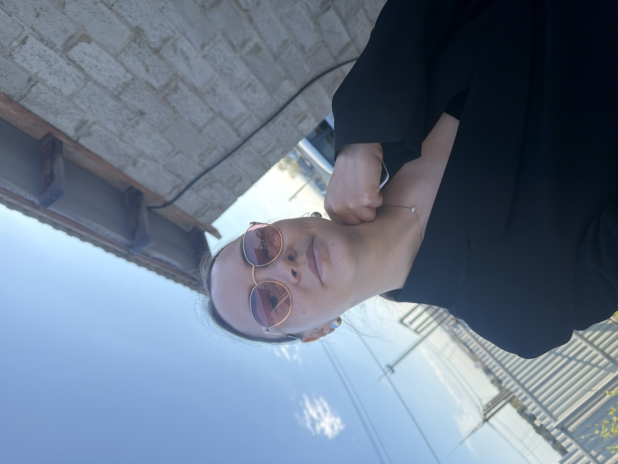 The image shows a person wearing rose-tinted sunglasses and a black blazer. The background includes a brick structure, blue sky with a cloud, and a metallic fence-like structure. Based on the location information provided, this scene takes place in the city of Chaikovsky, Russia.
