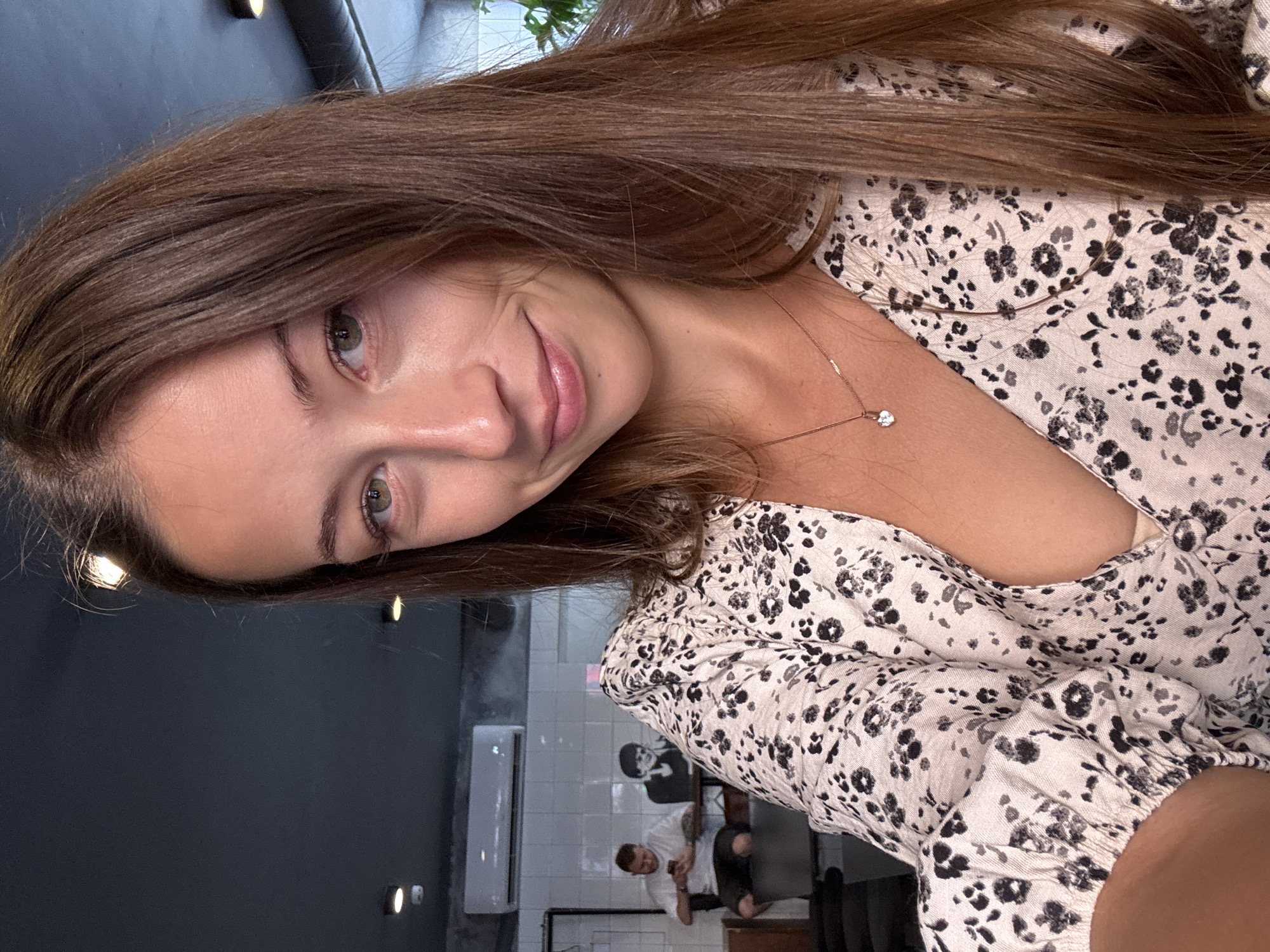 The image displays a selfie of a young adult female taken indoors, presumed to be within a cafe or restaurant setting. The female subject, positioned centrally and looking directly at the camera, has long, straight, brown hair, green eyes, and a slight, closed-mouth smile. She is wearing a light-colored V-neck blouse or dress with a dark floral pattern in black and grey, and a thin gold-colored necklace with a small, heart-shaped pendant is visible on her chest.
The background shows a dark grey ceiling equipped with multiple recessed spot lights. A white split-unit air conditioner is mounted on a light grey wall section above a white tiled wall. Two male figures are present in the mid-ground. To the left, a man with visible arm tattoos is seated, wearing a white t-shirt and dark shorts, engaged with a smartphone. To his right, a dark, stylized silhouette or cutout of a person with dark hair is affixed to the wall, positioned behind a dark table. No discernible signage or unique architectural features are present to specifically identify the location within Saint Petersburg, Russia.