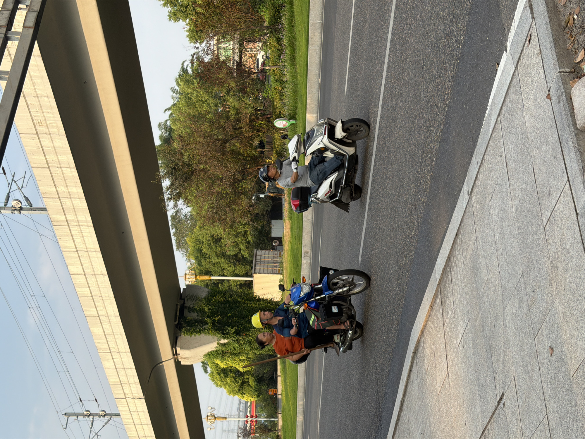 In Jiangjin, China, two motorcycles are captured in motion on a paved road under an elevated train track. On the left, a blue motorcycle carries three people: a driver, a woman in an orange shirt riding pillion with a yellow helmet, and another woman in an orange shirt riding behind her, holding a long wooden pole. On the right, a white and silver scooter is ridden by a man in a gray shirt and blue jeans. The road is bordered by a sidewalk on the left and a grassy area with trees and buildings on the right. The sky is clear and blue, suggesting it is daytime.