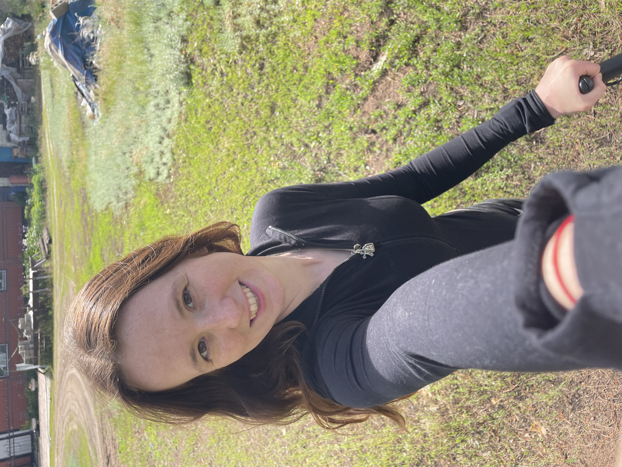 A smiling female, estimated to be in her 20s-30s, with long brown hair and a freckled complexion, takes a selfie outdoors in Vasilevka, Russia. She wears a black long-sleeved top with a partially visible zipper and a silver-colored pendant featuring two small stylized figures on a chain around her neck. A red string bracelet is present on her left wrist, and her right hand holds a black extension pole or similar device, directed towards the camera. The subject is positioned against a bright, sunlit backdrop of a grassy and unpaved area. The ground features a mix of green grass, lighter green low-lying vegetation, and exposed reddish-brown dirt, with a curved dirt path winding through the midground. In the background, a reddish-brick building with windows is visible to the left. To the right, various materials, including stacked wooden pallets and items covered by blue and grey tarps, are accumulated near a smaller structure with blue siding or a blue door. The scene portrays a semi-rural or rural outdoor environment under clear daylight conditions.