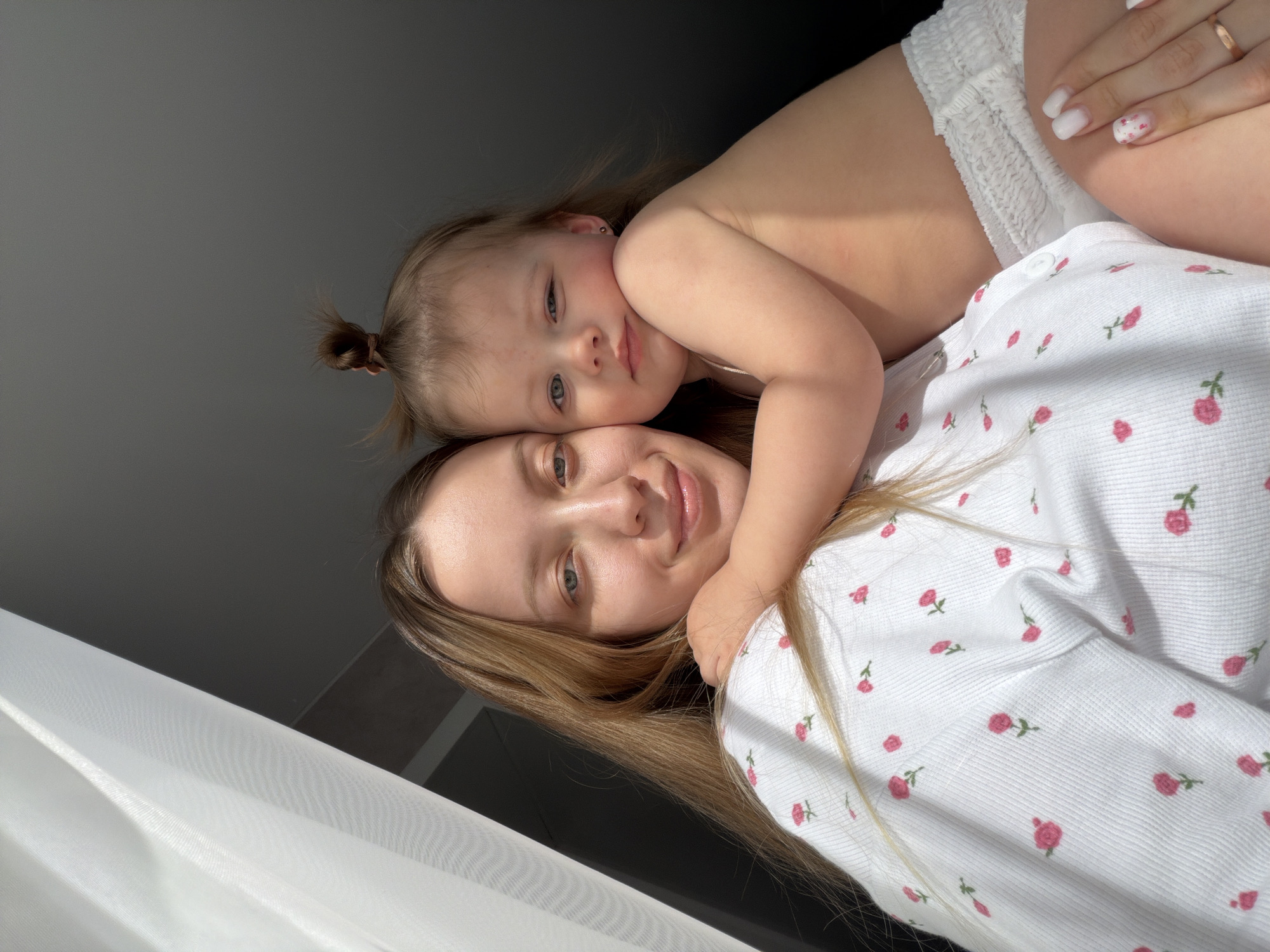 A woman with blonde hair is lying down and holding a toddler wearing a diaper. The woman is wearing a white shirt with small pink floral patterns. The toddler is lying on top of her, with their head resting on her shoulder. The background is a gray wall with a curtain in the bottom left corner. This image does not provide enough information to determine that the location is Reshetnikovo, Russia.