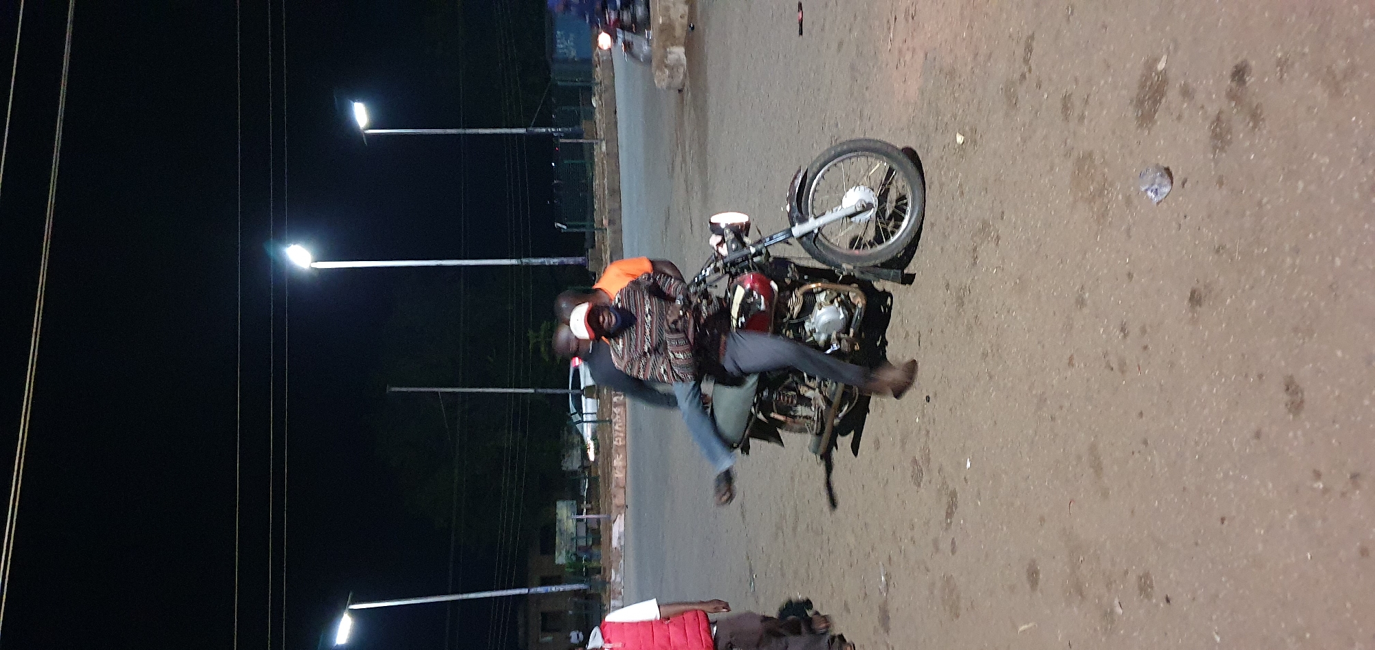 At night in an outdoor paved area in Bukuru, Nigeria, three men are visible. The central figure, wearing a patterned long-sleeved shirt and dark pants, is seated on a motorcycle, with his left leg extended backward. His face is partially obscured by a light-colored baseball cap and a dark facial covering. To his right, a man in an orange t-shirt stands with his hands on the motorcycle. Behind the seated man, another man in a dark shirt is also positioned on or near the motorcycle. To the left of the motorcycle, a person wearing a red vest over a white shirt and dark pants is standing. The environment is illuminated by several streetlights on poles, and power lines are visible overhead. The ground is a dusty, paved surface with some scattered debris. In the background, there are buildings and a car.