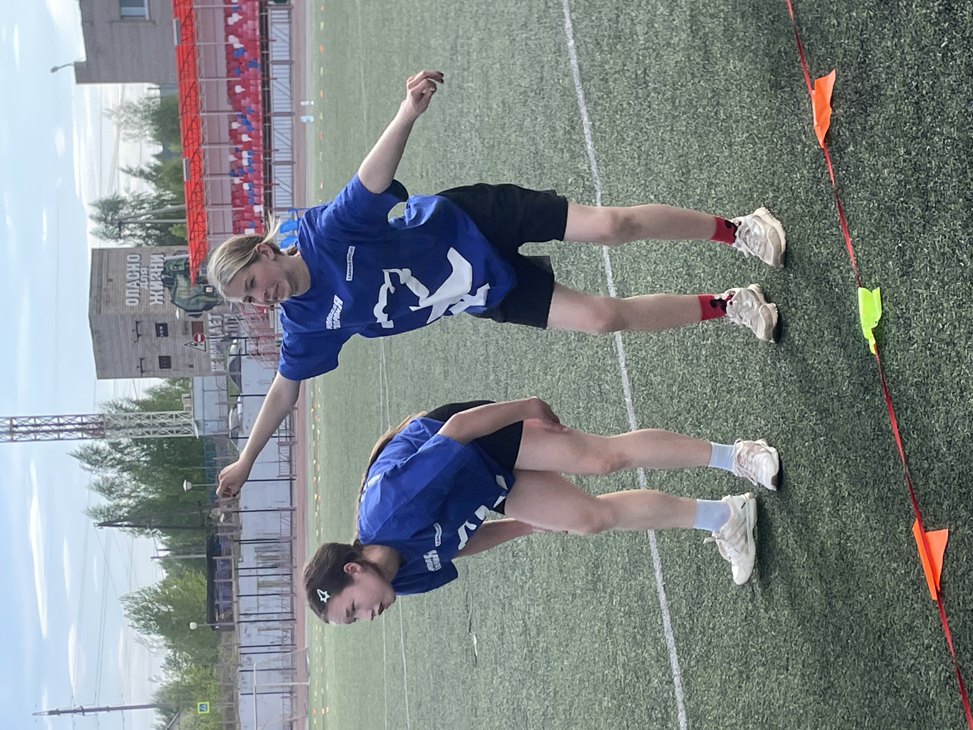 Two young women in matching blue T-shirts and black shorts stretch on a green athletic field marked with white lines. The woman on the left faces towards the bottom of the image and is bent over, with a white star clip in her dark hair. The other woman stands with her arms outstretched. They both wear white sneakers, and the woman on the top has red socks. A red barrier with orange and yellow flags runs along the right side of the frame. In the background, there's a building with the sign "ОПАСНО ДЛЯ ЖИЗНИ" (Dangerous for Life), red and white stadium seating, and trees. This is Zav'yalovo, Russia.