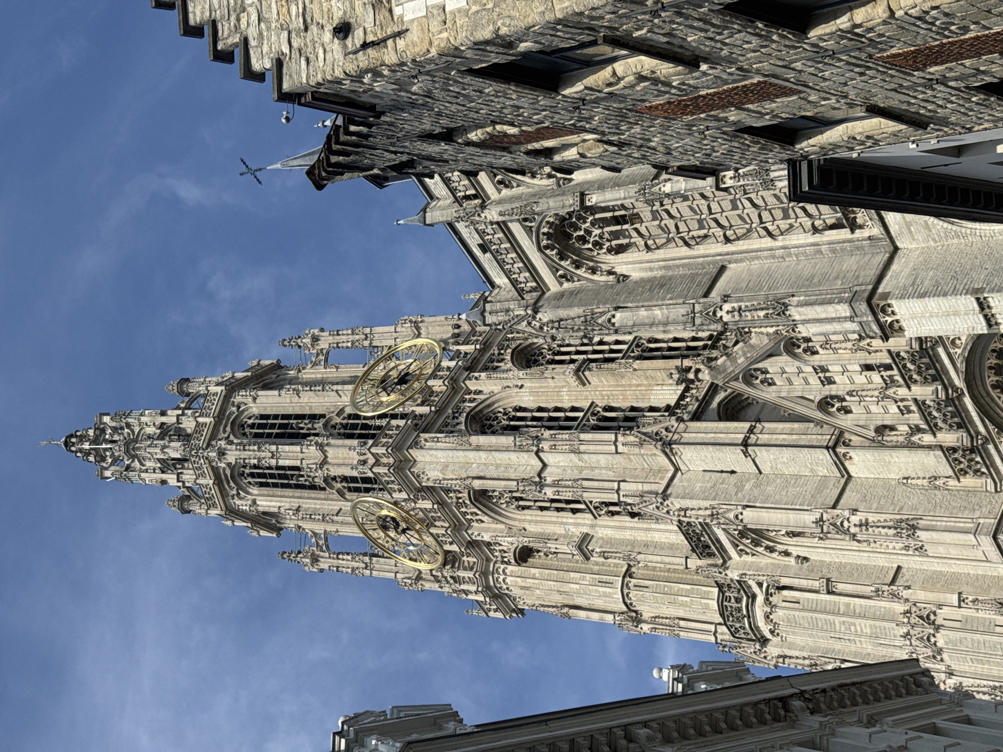 The image shows a low-angle view of the Cathedral of Our Lady in Antwerp, Belgium, on a clear day. The magnificent Gothic cathedral, with its tall, ornate spire and intricate stone carvings, dominates the frame. Two large, golden clock faces are visible on the tower. To the right of the cathedral, a stone building with a pointed roof and some visible windows juts into the frame. The sky is a vibrant blue with a few wispy clouds. The image is composed of sharp details, capturing the architectural grandeur of the cathedral and the texture of the stone building. There are no people or animals visible in the image.