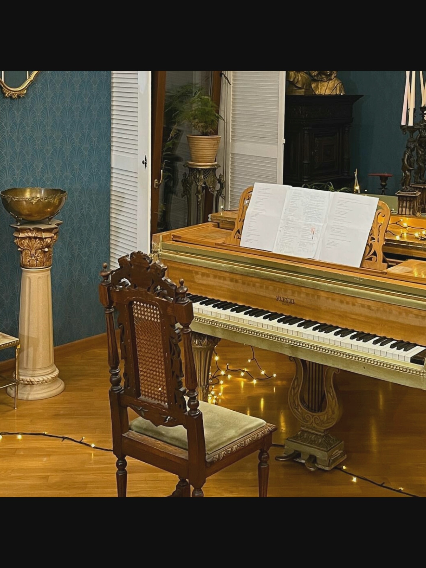 An indoor scene depicts a grand piano with sheet music on its stand. To the left of the piano is a wooden chair with a cane back and a green cushioned seat. A decorative pedestal with a golden bowl sits further to the left, against a teal patterned wallpaper. In the background, a plant in a pot rests on an ornate stand near a white louvered door. Behind the piano, a dark wooden cabinet with a bust on top is visible, along with candelabra holding unlit candles and other decorative figures. The floor is made of polished wood, and a string of small lights runs across it, illuminating the area around the piano and chair. The scene is well-lit, with the warm glow from the lights adding to the ambiance. There are no people visible in the image. The exact location in the city of Saint-Août, France is not provided.