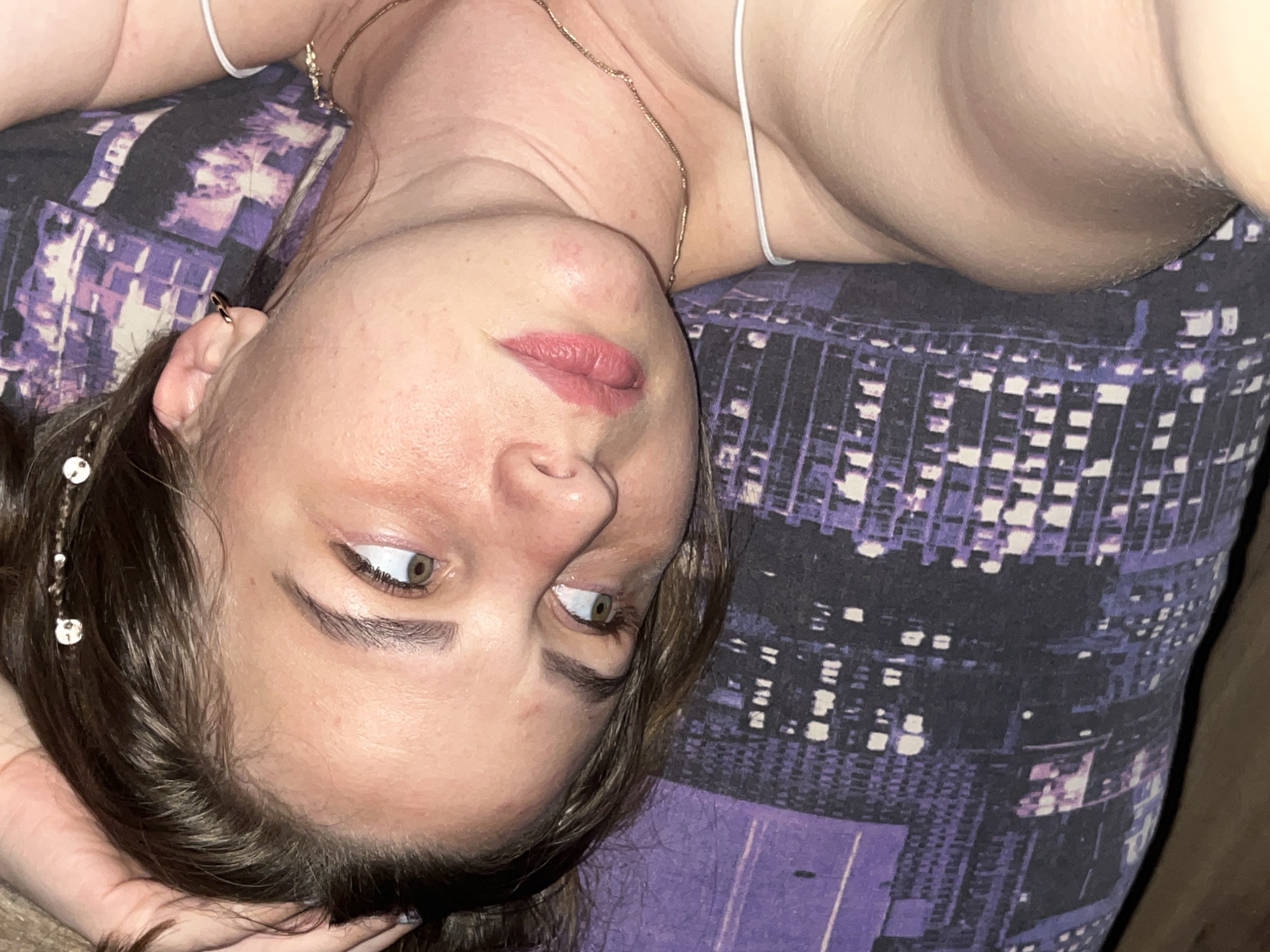An overhead shot shows a person, likely female, lying on their back in an indoor setting, with their head positioned towards the bottom left of the frame and their body extending upwards and to the right. The person has brown hair, styled with small, circular, reflective charms adorning a strand near the temple, and light green or hazel eyes that are open and directed upwards and to the side, not directly at the camera. Their lips are slightly parted, showing a soft pink lip color. A small, gold-colored hoop earring is visible in the person's left ear, and a delicate gold-toned chain necklace is worn around the neck. They are wearing a light-colored, thin-strapped top or dress.
The person is resting on a surface covered with a distinctive dark purple and white patterned fabric, which features a repeating geometric or stylized cityscape design. A hand is partially visible, appearing to support the head or neck. The lighting in the image is soft and ambient, characteristic of an indoor setting, possibly in the evening or at night. The pose and setting suggest a moment of relaxation or contemplation in Izhevsk, Russia. No text is visible in the media file.