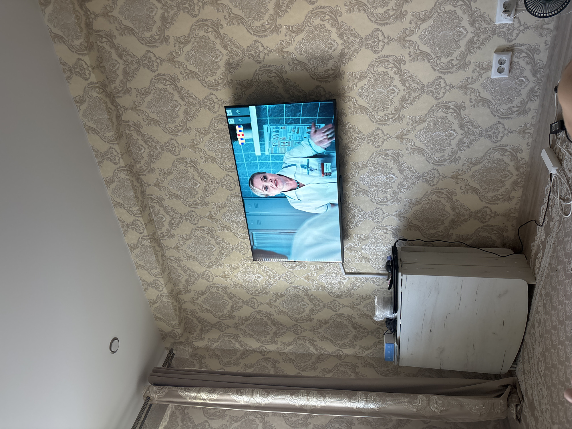 The image shows a wall-mounted television displaying a program. A woman in medical attire is visible on the screen. The wall is covered in patterned wallpaper. To the left of the television, a beige curtain hangs. Below the television, a white folding table is placed against the wall. Various objects, including a laptop, a mouse, and some cables, are on top of the table. The floor is covered with a patterned rug. Electrical outlets are visible on the wall. The scene appears to be an indoor residential setting, possibly a living room, in the city of Nefteyugansk, Russia.