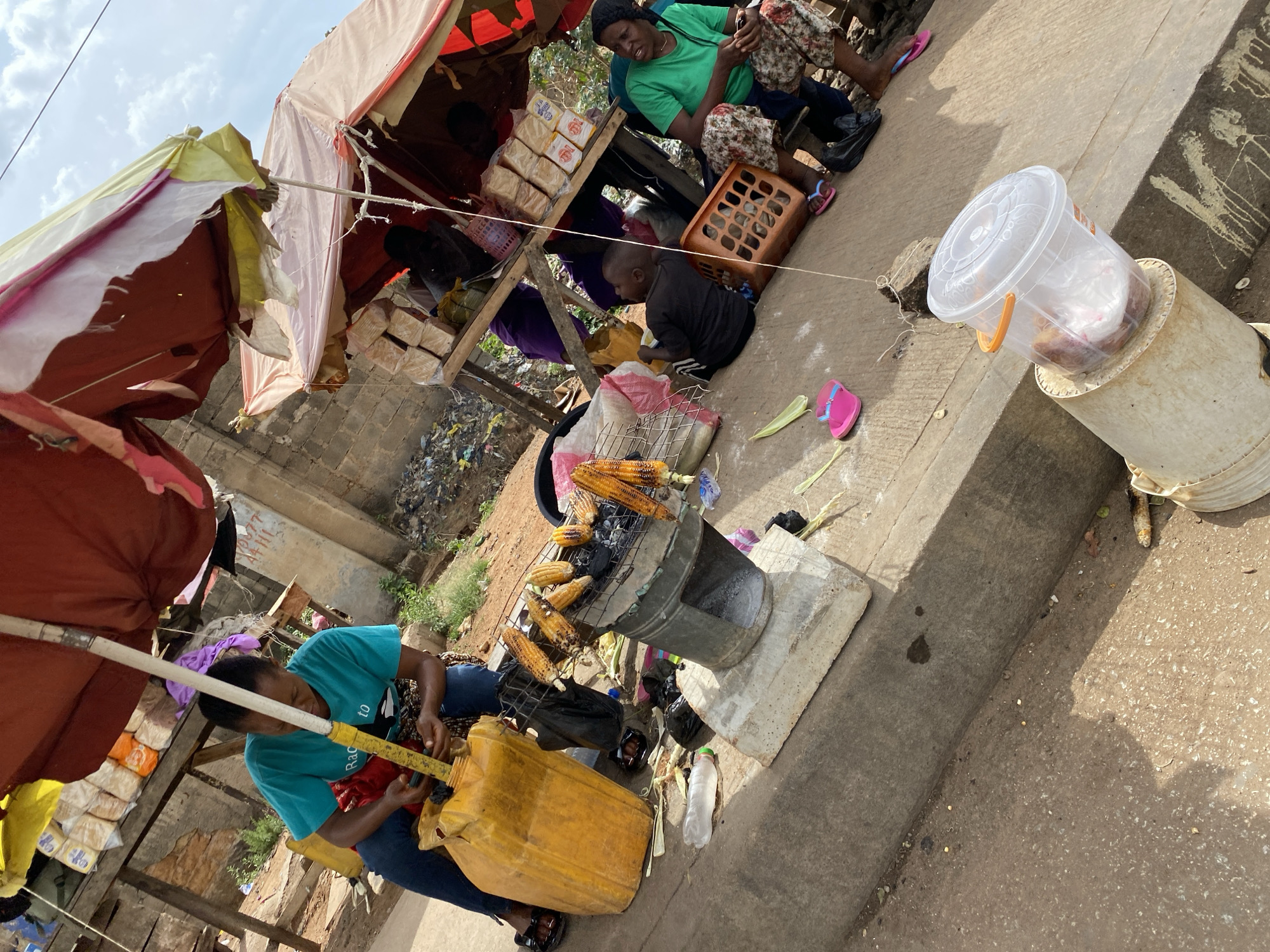 This image captures a vibrant roadside market scene in Bukuru, Nigeria, during the daytime under a clear, bright sky.
In the foreground, a woman wearing a teal t-shirt and blue jeans is actively preparing items, possibly food or drink, from a large yellow plastic jerrycan. Next to her, a makeshift charcoal grill holds several ears of roasted corn, indicating a food stall. The ground is concrete, with some corn husks and debris scattered around the grill.
Behind the corn vendor, two makeshift market stalls are set up under colorful fabric awnings, providing shade. One stall displays stacks of packaged loaves of bread. Another woman, dressed in a green t-shirt and patterned skirt, sits near this stall, looking down at something in her hands. A young child in a dark shirt sits on the ground nearby. Further into the background, what appears to be discarded trash is visible.
Other notable objects include a clear plastic bucket with a lid containing food items, resting on a pipe, and discarded plastic bottles. The woman in the teal shirt has partially visible text on her shirt that reads "Racetr...". The overall atmosphere suggests a busy, informal commercial area.