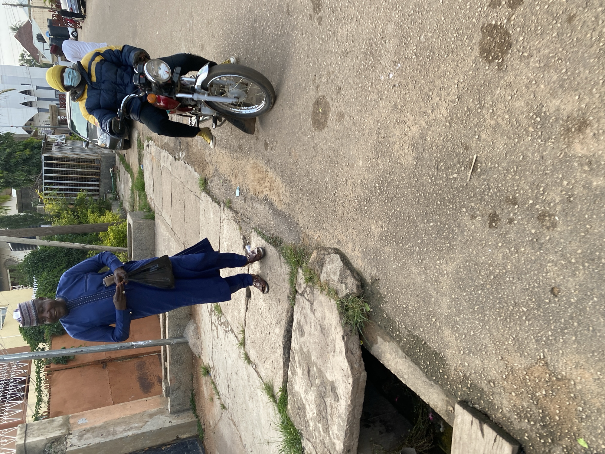 The image depicts a street scene in Jos, Nigeria. In the foreground, a man in a blue traditional outfit and a patterned cap walks on a paved walkway next to a drainage ditch. He is holding a black plastic bag in his left hand. Behind him, a person wearing a yellow beanie, a blue and yellow jacket, black pants, and yellow shoes rides a red motorcycle. This person is also wearing a light blue face mask. The street is asphalt, and there are buildings and vegetation visible in the background, along with parked cars. The overall environment suggests an urban setting, likely in Jos, given the context provided.