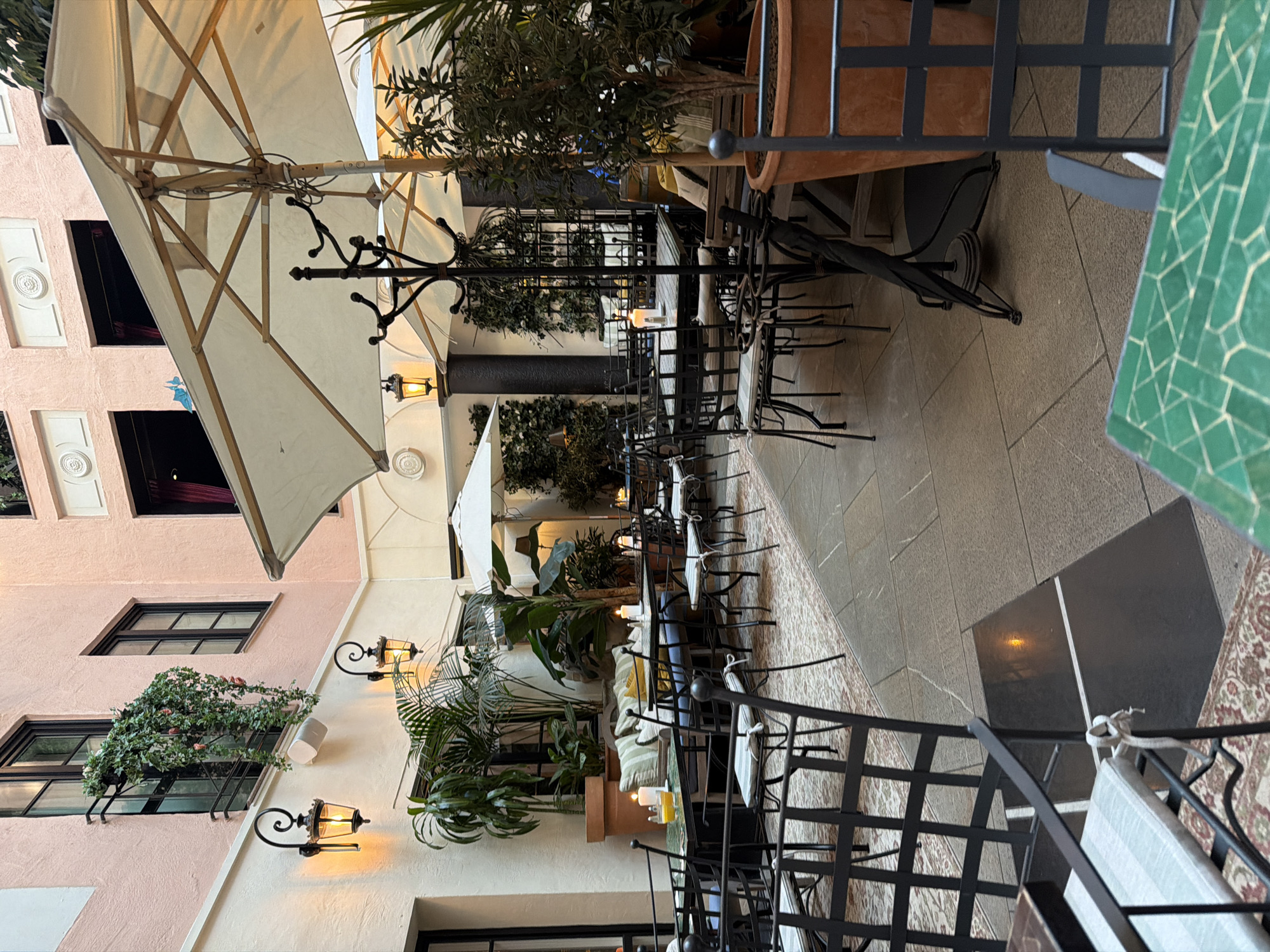 The image depicts an outdoor seating area of a restaurant or cafe, likely in Malmö, Sweden, given the query. The scene is an enclosed courtyard with a pale pink stucco building surrounding it. There are several tables and chairs set up for dining.
Dominating the upper right of the frame is a large, off-white patio umbrella with a wooden frame. In the foreground, black metal chairs with striped cushions surround tables. Some tables have candles lit on them.
The courtyard is decorated with numerous potted plants and hanging greenery, creating a lush atmosphere. Two ornate black wall lamps with yellow light bulbs are visible on the building's exterior, casting a warm glow. The floor is paved with stone tiles, and a patterned rug is visible beneath some of the tables and chairs.
The architecture of the building includes dark-framed windows and decorative circular elements above some of them. The overall impression is of a serene and inviting outdoor dining space. No people are visible in the image.
