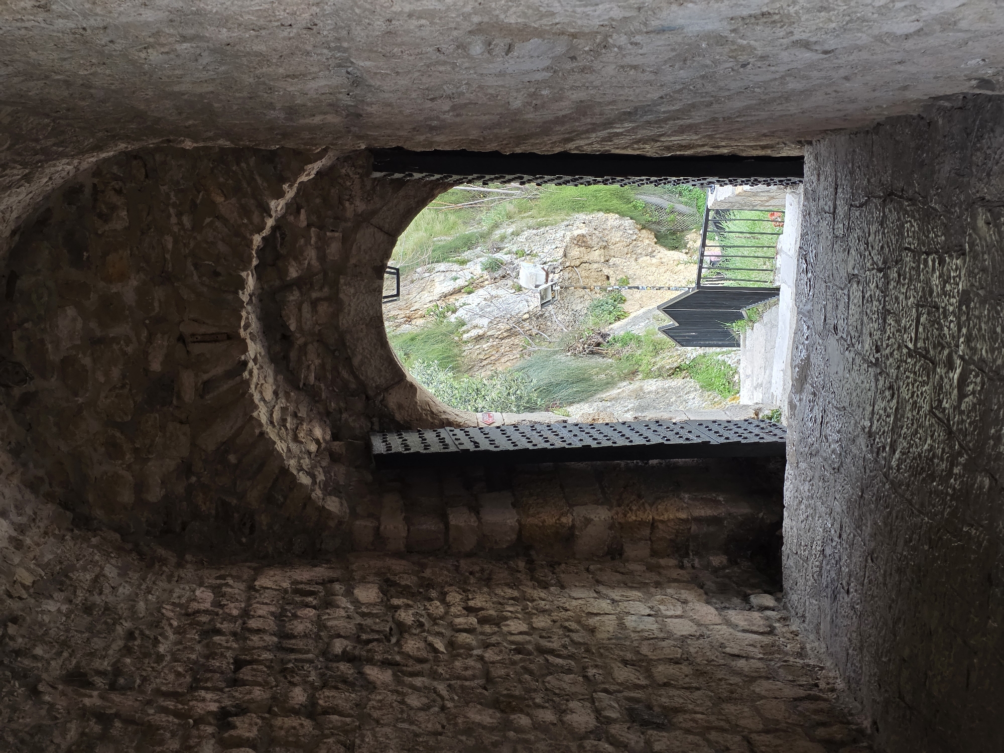 The image presents a view looking outwards from a dimly lit, vaulted stone archway, identified as being in Ulcinj - Ulqin, Montenegro. The interior walls and arched ceiling of the passage are constructed from rough-hewn, irregular brown and grey stones. The floor is paved with dark, uneven flagstones or cobblestones, exhibiting some reflective properties.
At the archway's exit, a heavy, dark gate, possibly metal and featuring a studded surface, is partially open, with its right panel angled inward. Beyond the archway, a steep, brightly illuminated rocky incline dominates the background. This natural rock face is covered with green grass, shrubs, and scattered dry branches. A light-colored streetlamp or floodlight on a pole is visible against the rocky terrain to the left. Immediately outside the archway, a paved path or set of steps descends, flanked on the left by a dark, possibly black, metal railing with a distinctive zig-zag or pointed design along its top edge. No human figures or discernible activity are present.