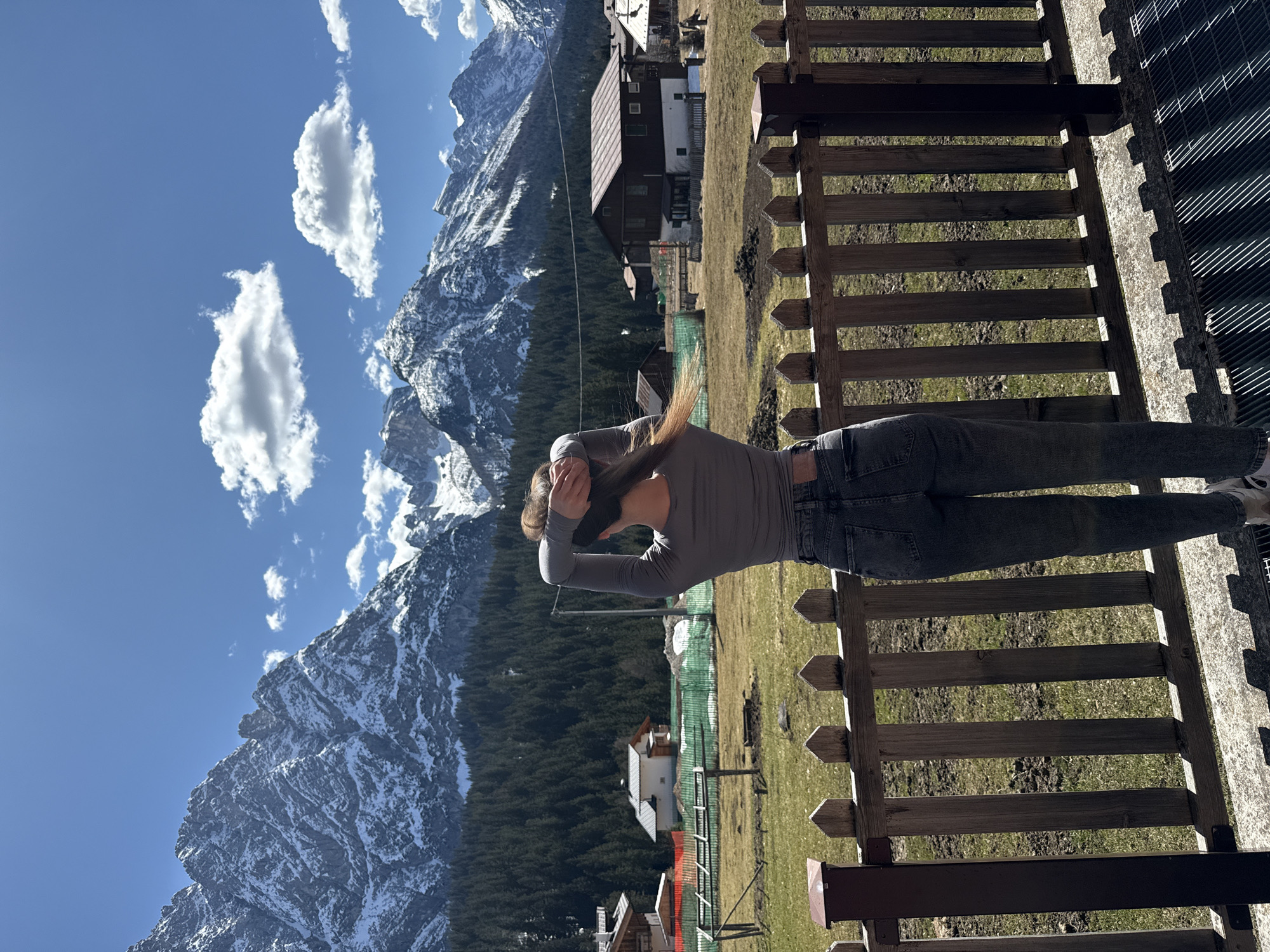 A landscape photograph taken in Sappada, Italy, shows a person with their back to the camera, facing a mountain range. The person is standing behind a wooden fence, wearing a gray long-sleeved shirt and dark jeans, with their hands behind their head. The mountains in the background are partially covered in snow and surrounded by a dense forest. The sky is blue with scattered white clouds. In the midground, there are buildings and structures typical of the Sappada area. The foreground includes a fence and some grassy terrain. The perspective is slightly angled, giving a panoramic view of the scene.