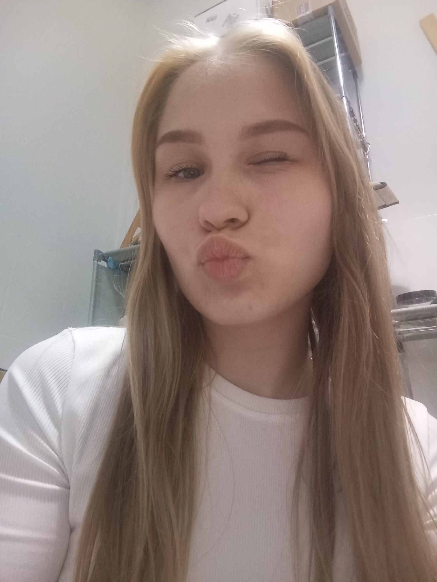 A young woman with light blonde hair, wearing a white ribbed long-sleeved shirt, is posing for a selfie. She is winking her left eye and puckering her lips into a kiss. The background is a plain white wall. A gray laundry basket and a metal shelf unit are partially visible. Based on the objects visible in the background, the photo may have been taken indoors, possibly in a utility or storage area. There is no information in the image to indicate that it was taken in Krasnodar, Russia.