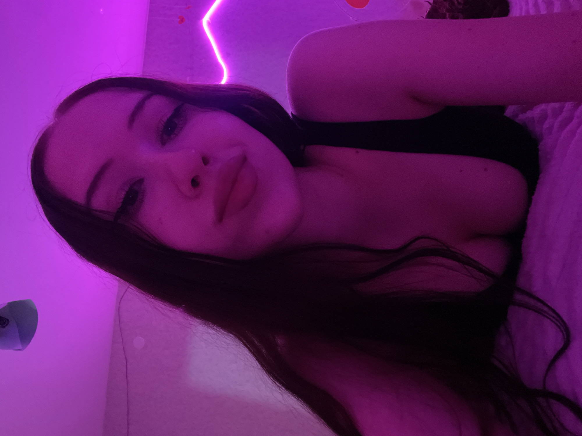 The image is a close-up, low-angle selfie of a young woman with long, dark hair, lying down in what appears to be a bedroom. She is wearing a black top that reveals part of her cleavage. The room is illuminated with pink and purple lighting, possibly from LED strip lights which are visible along the wall in the background. There is also a stuffed animal hanging from the ceiling. The woman's facial expression is relaxed. The scene is not identifiable as being located in Votkinsk, Russia.