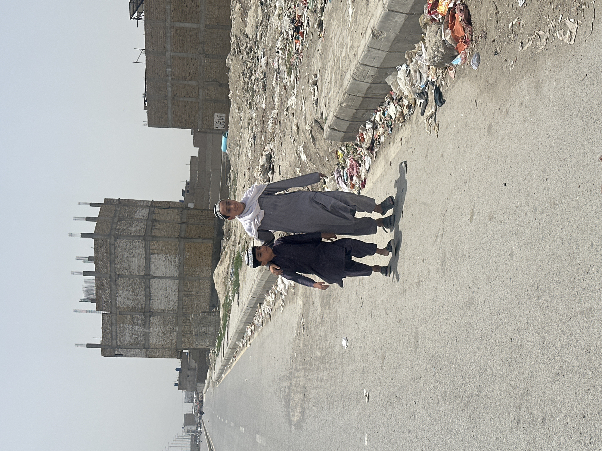 Two individuals, likely males, are positioned on the right-hand side of a road within Quetta City Tehsil, Pakistan. The older of the two is attired in a gray tunic and a white kufi. The younger male is wearing a black outfit and a similar, dark-colored kufi. Both are standing adjacent to a curb that abuts a landfill or a heavily littered area. The roadway is paved, and the perspective suggests a straight stretch of road leading to a distant horizon with visible traffic. On the opposite side of the street, there are buildings under construction.