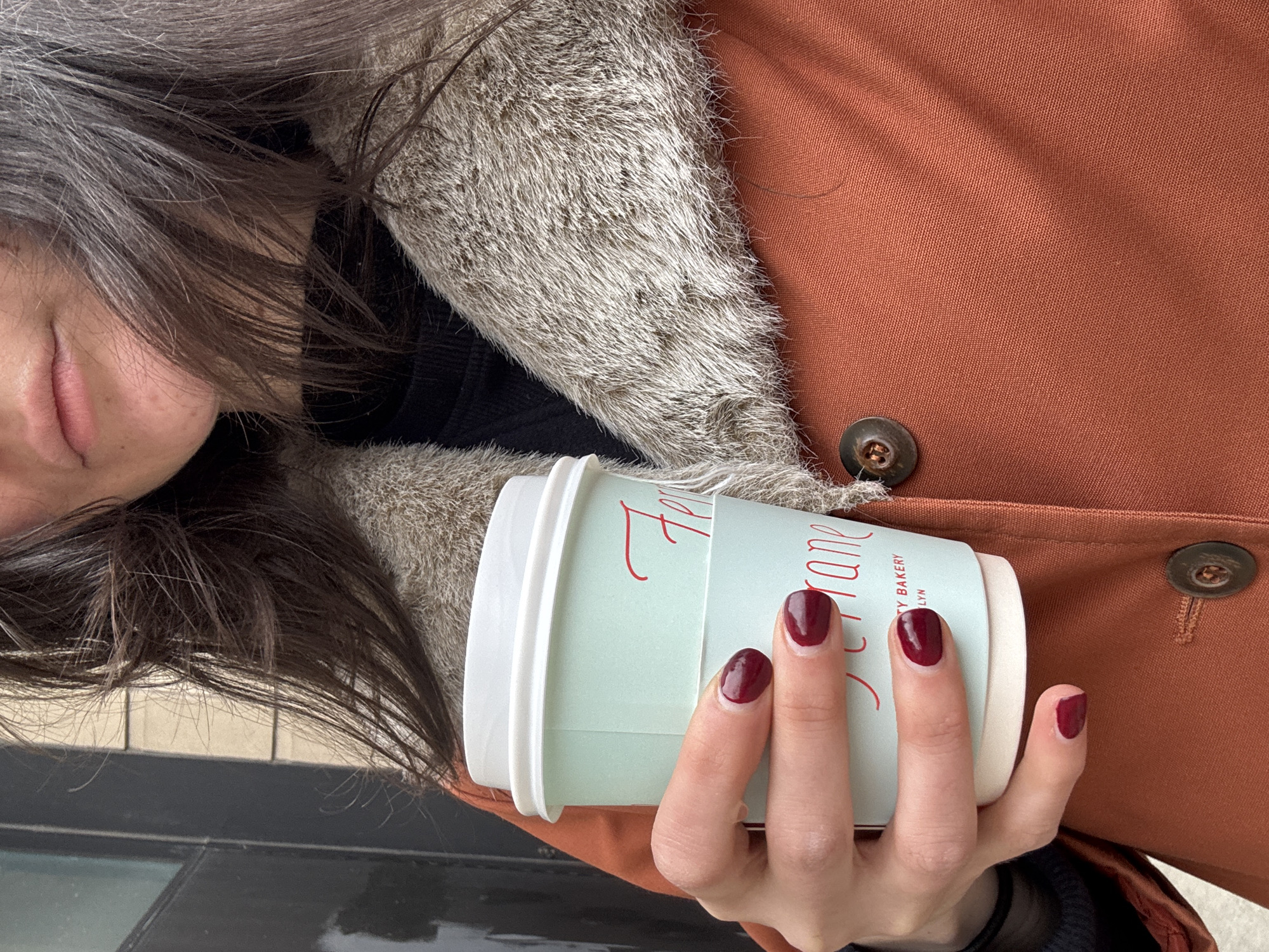A woman's partial face and hand holding a Ferarane Bakery coffee cup in New York City, United States, are the focus of this shot. Her dark hair with gray streaks contrasts with the light tan furry collar of her jacket. The jacket is a rust-colored material with visible buttons. Her nails are painted a dark maroon color. A black shirt can be seen underneath the jacket. The coffee cup is a light green with the bakery name in red script. The background is a dark, indistinct surface.
