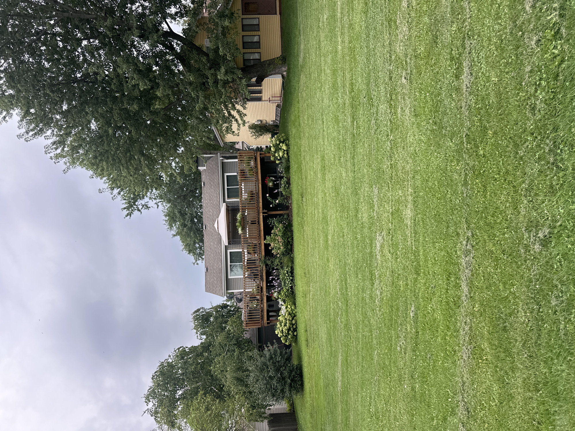 A backyard scene in Streamwood, United States of America, features a large, freshly mown lawn in the foreground. Behind the lawn, a multi-level wooden deck extends from the back of a two-story house. The house is gray with a brown roof and features several windows. On the deck, there is outdoor furniture and a white gazebo. To the right of the gray house, a smaller, yellow house is visible, partially shaded by a large tree. The sky is overcast and cloudy. The overall impression is a suburban residential setting on a cloudy day.