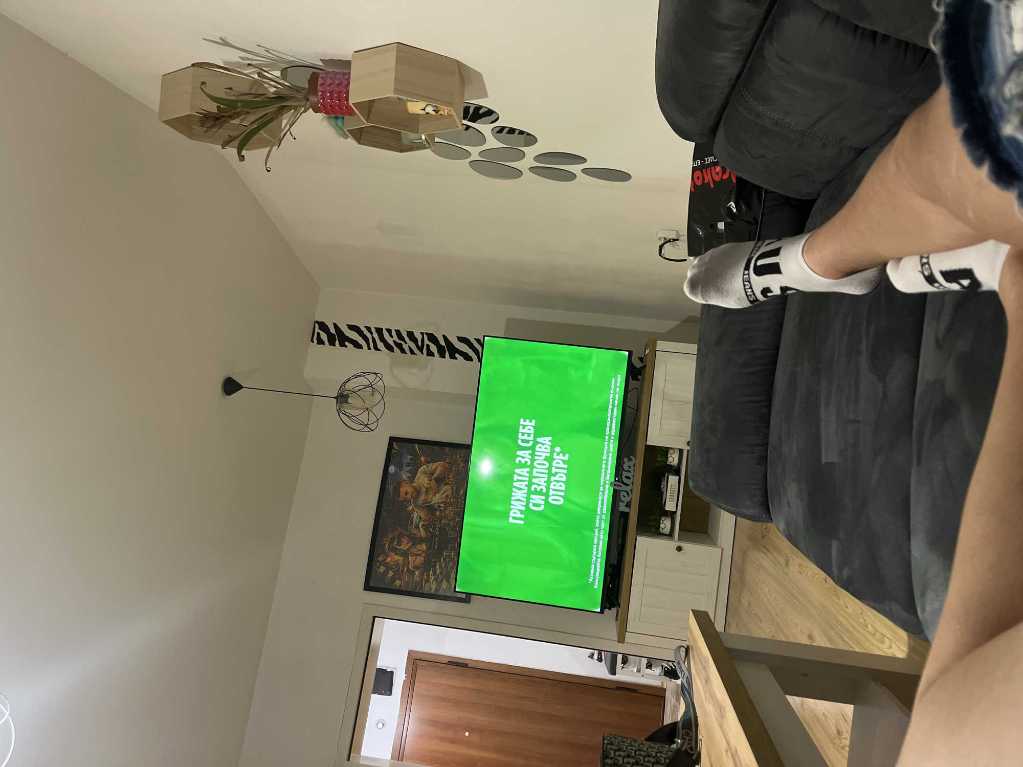 The image captures an indoor scene, likely a living room or lounge area in Plovdiv, Bulgaria, during the evening or night. The perspective is from a person relaxing on a dark gray upholstered sofa, with their lower legs and feet visible in the foreground. The person is wearing white ankle socks with black lettering, one clearly showing "JEANS" and another partial logo. A black bag with "kako" and Greek text "ΓΙΔΕΣ - ΕΠΙ" is partially visible next to the sofa.
A flat-screen television dominates the center, displaying a bright green background with white Bulgarian text. The primary message reads: "ГРИЖАТА ЗА СЕБЕ СИ ЗАПОЧВА ОТВЪТРЕ*" (CARE FOR YOURSELF STARTS FROM WITHIN*). Smaller text below translates to: "Activita contains calcium, which contributes to the normal function of digestive enzymes. Consume Activita as part of a varied and balanced diet and a healthy lifestyle." The word "relax" is also displayed vertically in a cursive font.
Below the television, a light-colored wooden cabinet provides a base, while above it, a small shelf holds decorative items, including a box labeled "SERVETT". To the left of the TV, a framed artwork depicting figures with money is mounted on the wall. A section of the wall behind the TV features a decorative black and white zebra-stripe pattern.
The room's ceiling is adorned with a unique decorative feature consisting of wooden hexagonal boxes containing plants and small circular mirrors arranged in a cluster. A contemporary ceiling light fixture with a wireframe design hangs in the mid-ground. The room is characterized by light-colored walls and a wooden floor, creating a modern and comfortable ambiance. A closed wooden door is visible in the background, suggesting an entryway.