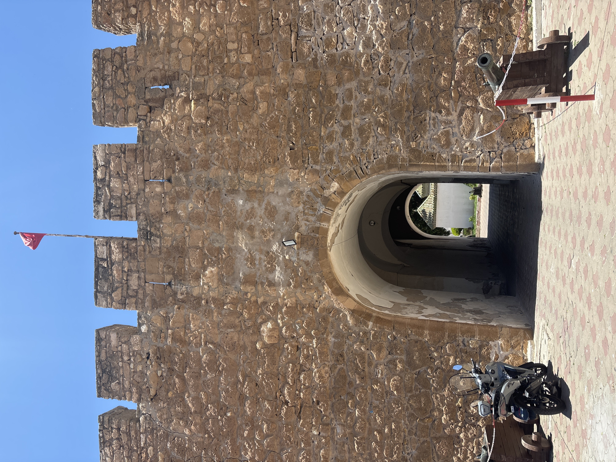 A full exterior shot captures a historical stone fortification on a sunny day. The fortification's thick, rough-hewn stone walls are topped with crenellations. A red flag flies from a flagpole positioned centrally on the battlements. An arched gateway, leading into a dimly lit passage, pierces the wall. To the left of the gateway, a light-colored scooter is parked. On either side of the gateway, antique cannons are displayed on wooden carriages. The ground in front of the fortification is paved with patterned tiles. The sky above is a clear, bright blue.