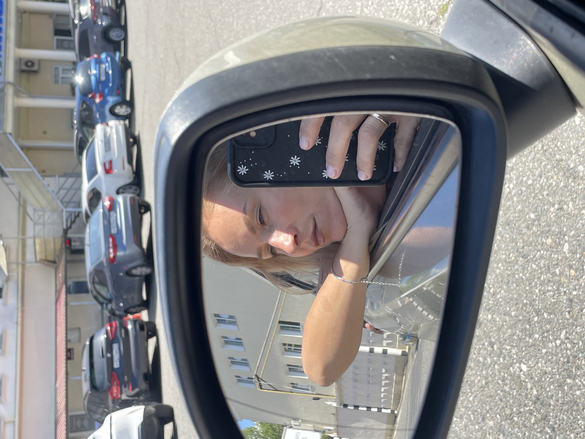 The image is a selfie taken in the side mirror of a car. A woman is seen in the reflection, holding a phone with a white flower-patterned black case. The woman has light skin and blonde hair. She is wearing a ring on her left ring finger and a bracelet on her left wrist. The car is parked next to a multi-story building with white exterior walls and multiple windows. Several cars are parked in a line behind the car. Based on the architectural style and the Cyrillic text visible in the reflection, the location is likely in Sochi, Russia.