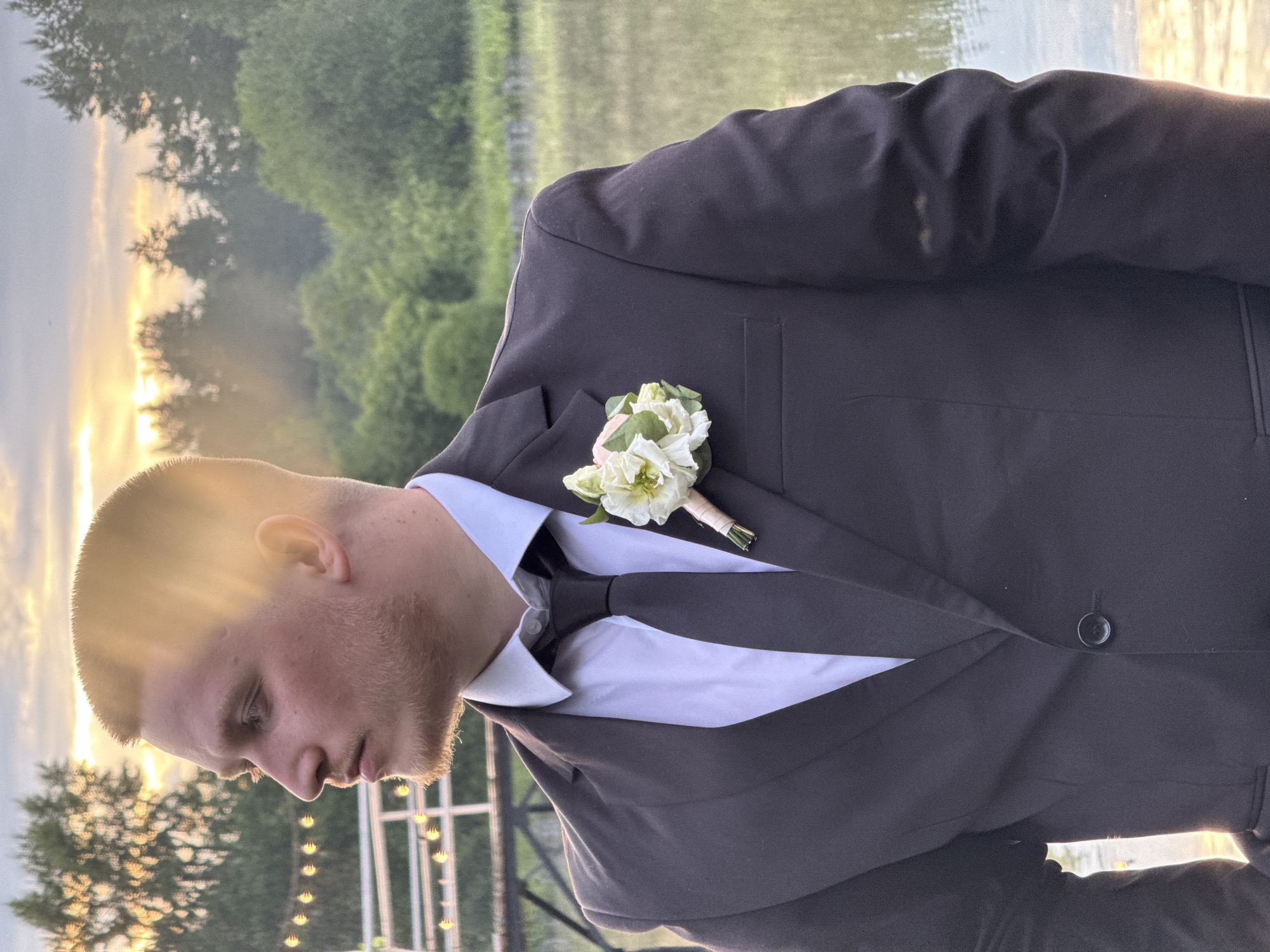 This media file is a close-up, rotated portrait of a young man dressed in a dark suit, white shirt, and dark tie. He has a boutonnière consisting of light-colored flowers and greenery pinned to his lapel. The man has short hair and is looking downwards, with a neutral expression. Sunlight is casting a strong, golden glow on his face and hair, suggesting it is either sunrise or sunset.
The background indicates an outdoor setting with a body of water in the distance and trees lining the shore. There are also string lights visible, possibly indicating an event or celebration. The overall atmosphere is serene, enhanced by the warm, soft lighting. The location context provided is Votkinsk, Russia.