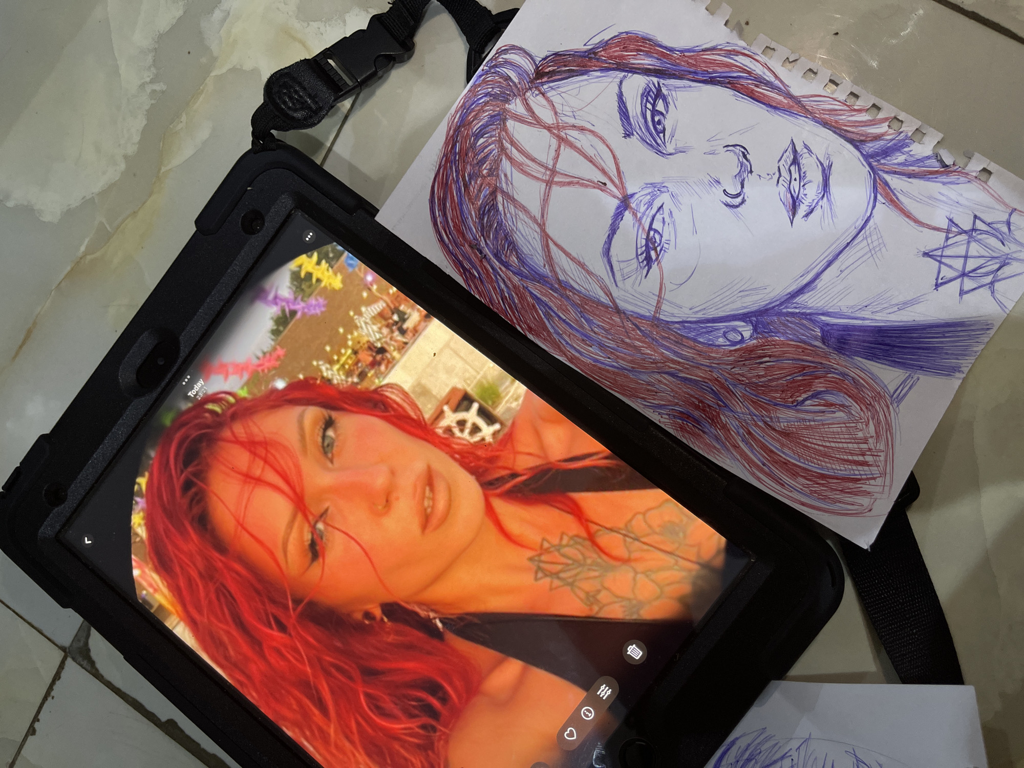 A close-up overhead shot shows a tablet displaying a selfie of a woman with bright red hair and prominent eye makeup. Next to the tablet, on a beige, patterned surface, lies a sketch of the same woman, rendered in blue and red ink. The sketch captures her face, neck, and chest, including a star-like tattoo. The tablet is encased in a black protective cover with a black strap attached. The overall impression is one of creative work in progress, with the digital image serving as a reference for the hand-drawn portrait. The scene does not provide any visual cues or context that would allow for the identification of the city of Abuja, Nigeria.