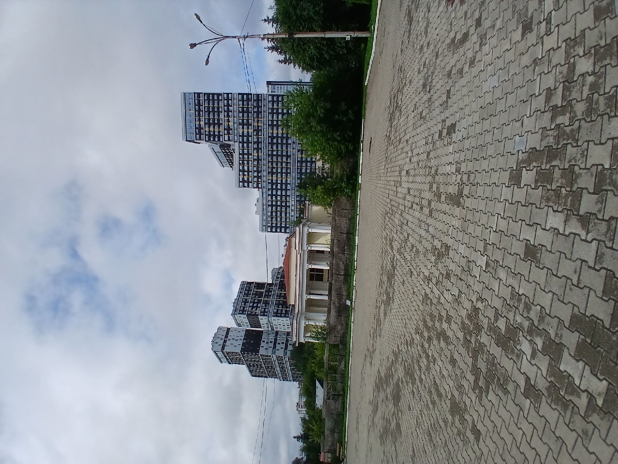 The image depicts a view of modern apartment buildings with a classical structure in the foreground, situated in Yekaterinburg, Russia. The foreground is an empty, wet paved area made of interlocking grey bricks. Behind this area, a light-yellow classical building with white columns and a red roof is visible. Further back, several tall, multi-story apartment buildings are seen with a distinctive tiered design, featuring dark facades with numerous windows. The sky is overcast with white and grey clouds. Trees and greenery are present between the classical building and the apartment complexes, as well as to the right of the image, where a street lamp with three lights stands. The scene suggests an urban environment on a cloudy day.