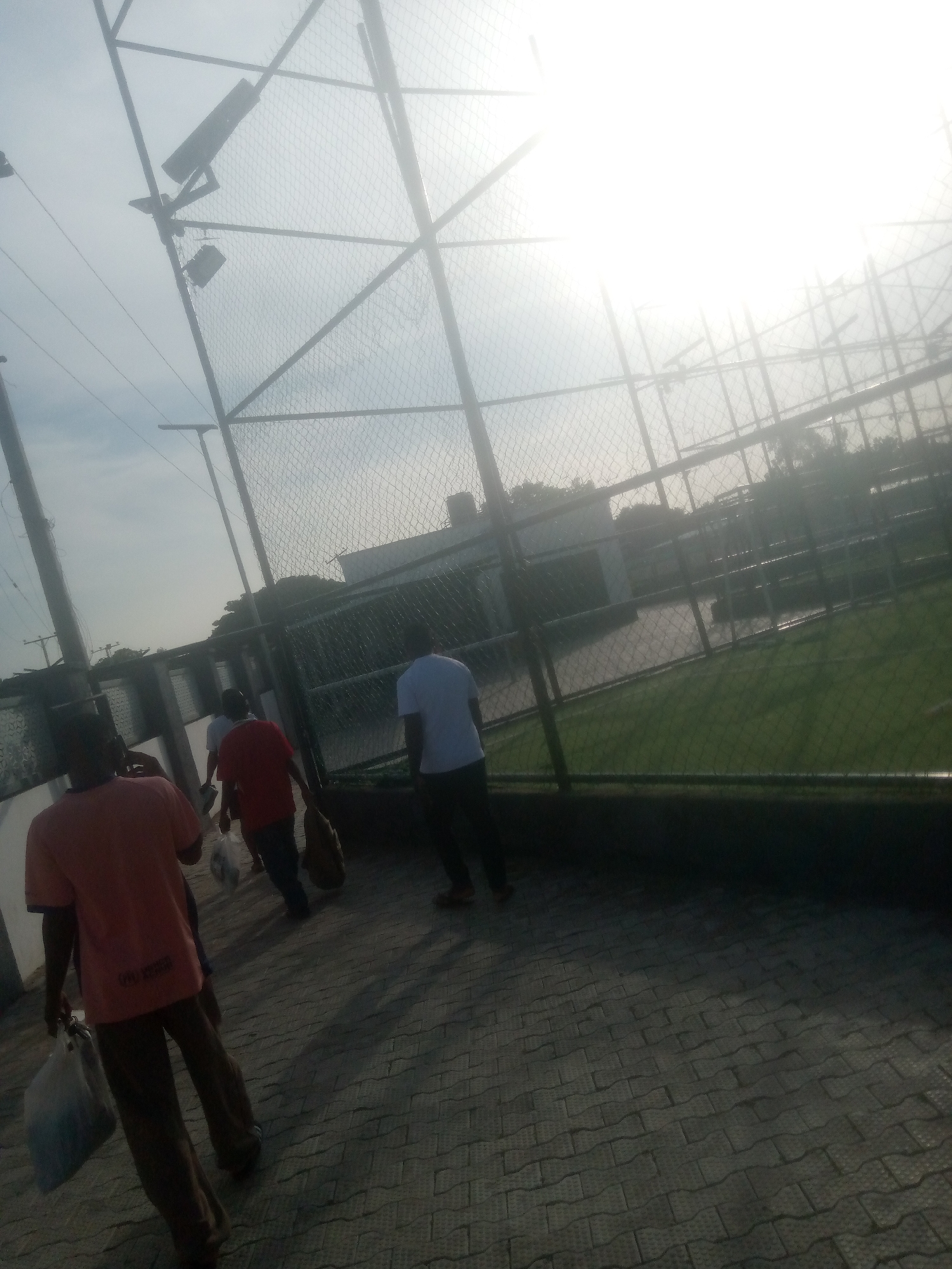 The image depicts a scene in Jalingo, Nigeria, with several individuals walking along a paved pathway adjacent to a fenced grassy area, possibly a sports field. The sun is bright and high in the sky, suggesting it is daytime, likely mid-morning or afternoon. The weather appears clear, with a bright, hazy sky due to the sun's intensity.
In the foreground, a person wearing an orange t-shirt and brown pants walks away from the camera, carrying a light blue plastic bag in their left hand. Behind them, another individual in a red t-shirt walks ahead, also carrying a bag. Further back, a person in a white t-shirt and dark pants walks, and another person is partially visible in the distance. The people appear to be casually dressed, suggesting a relaxed outing or activity.
The setting includes a large metal fence surrounding a green area, which is illuminated by the bright sunlight. Overhead, there are lights mounted on poles, indicative of a sports facility or an area used after dark. The pathway is made of interlocking pavers. The overall mood seems ordinary, with people going about their day. There is no visible text in the image.