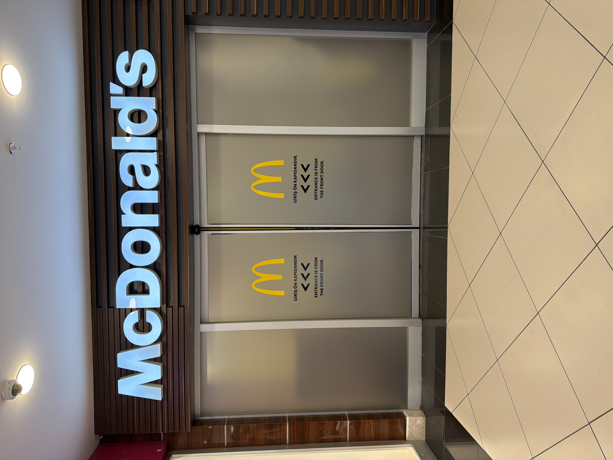 The image shows the entrance to a McDonald's restaurant. The sign "McDonald's" is prominently displayed vertically along the left side of the entrance. The entrance doors are frosted glass with the McDonald's logo and the text "GİRİŞ ÖN KAPIDANDIR" and "ENTRANCE IS FROM THE FRONT DOOR" printed on them in both Turkish and English. The floor in front of the entrance is tiled. Based on the multilingual signage (Turkish and English), this likely indicates that this McDonald's is located in Alanya, Turkey.