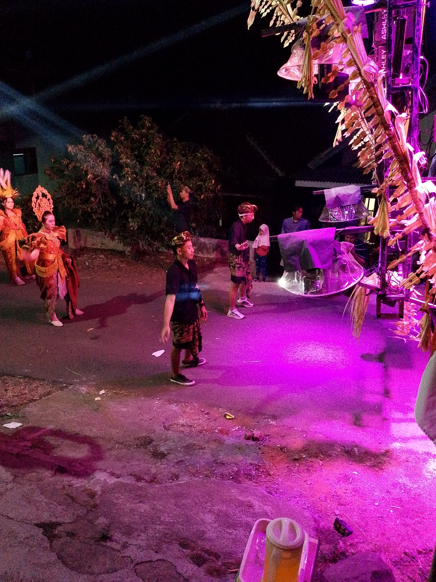 In the city of Babadan, Indonesia, at night, individuals are gathered outdoors for what appears to be a performance or event illuminated by purple and white stage lighting. In the foreground, on the right, a person in a black shirt and patterned shorts stands facing left, with a pink-rimmed container with a yellow liquid visible in the bottom right corner. To the left, two individuals in ornate, traditional costumes, featuring gold headdresses and vibrant fabrics, are positioned on an asphalt surface. Further back, several other people are visible, including one person in a black shirt observing the scene and another person in a blue shirt. Behind the individuals, trees and a building with a dark roof are present. A large speaker system is prominent on the right side of the image, adorned with foliage. The ground is a mixture of asphalt and dirt, with scattered debris and shadows cast by the lighting.