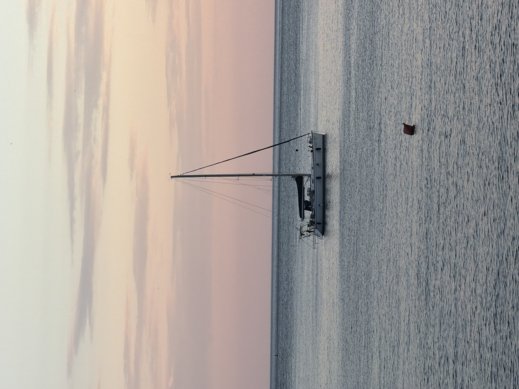The image shows a sailboat on calm water under a sky with pastel-colored clouds. To the right of the boat is a small red buoy.