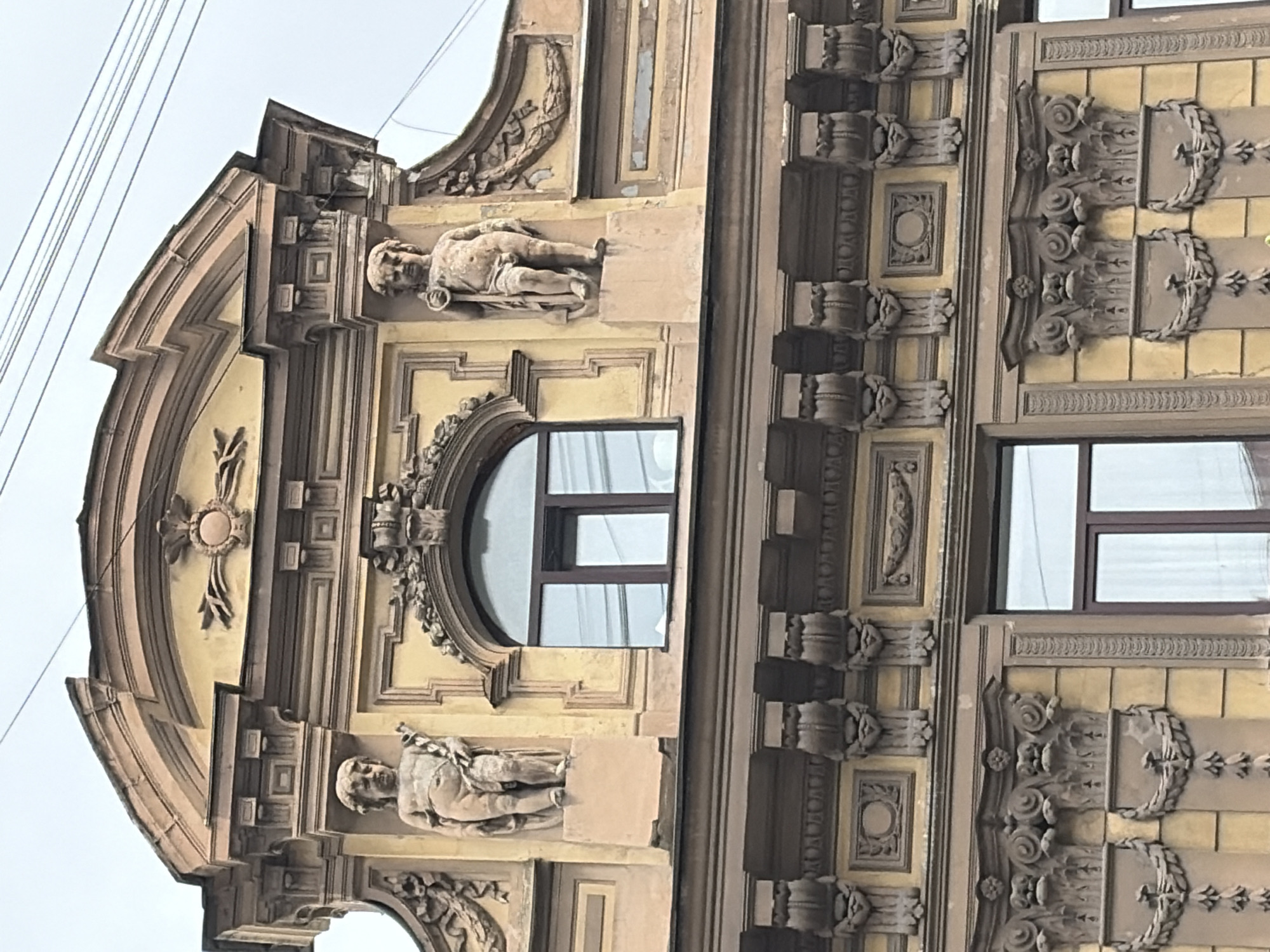 This is a medium-shot, eye-level, exterior photograph of the upper facade of a historic building in Saint Petersburg, Russia. The building features ornate architectural details, including two stucco statues of cherubic figures positioned symmetrically on either side of a large, arched window. The statues, made of a light-colored stone, appear to be holding staffs. The window has a dark brown frame and is divided into two panes. Above the window, a decorative medallion with a sunburst motif is visible. Below this level, another row of detailed ornamentation includes corbels and garlands. The facade is painted a pale yellow with contrasting dark brown decorative elements. Power lines are visible in the upper right corner against a cloudy sky. The overall style suggests pre-20th century architecture, common in the historic center of Saint Petersburg.