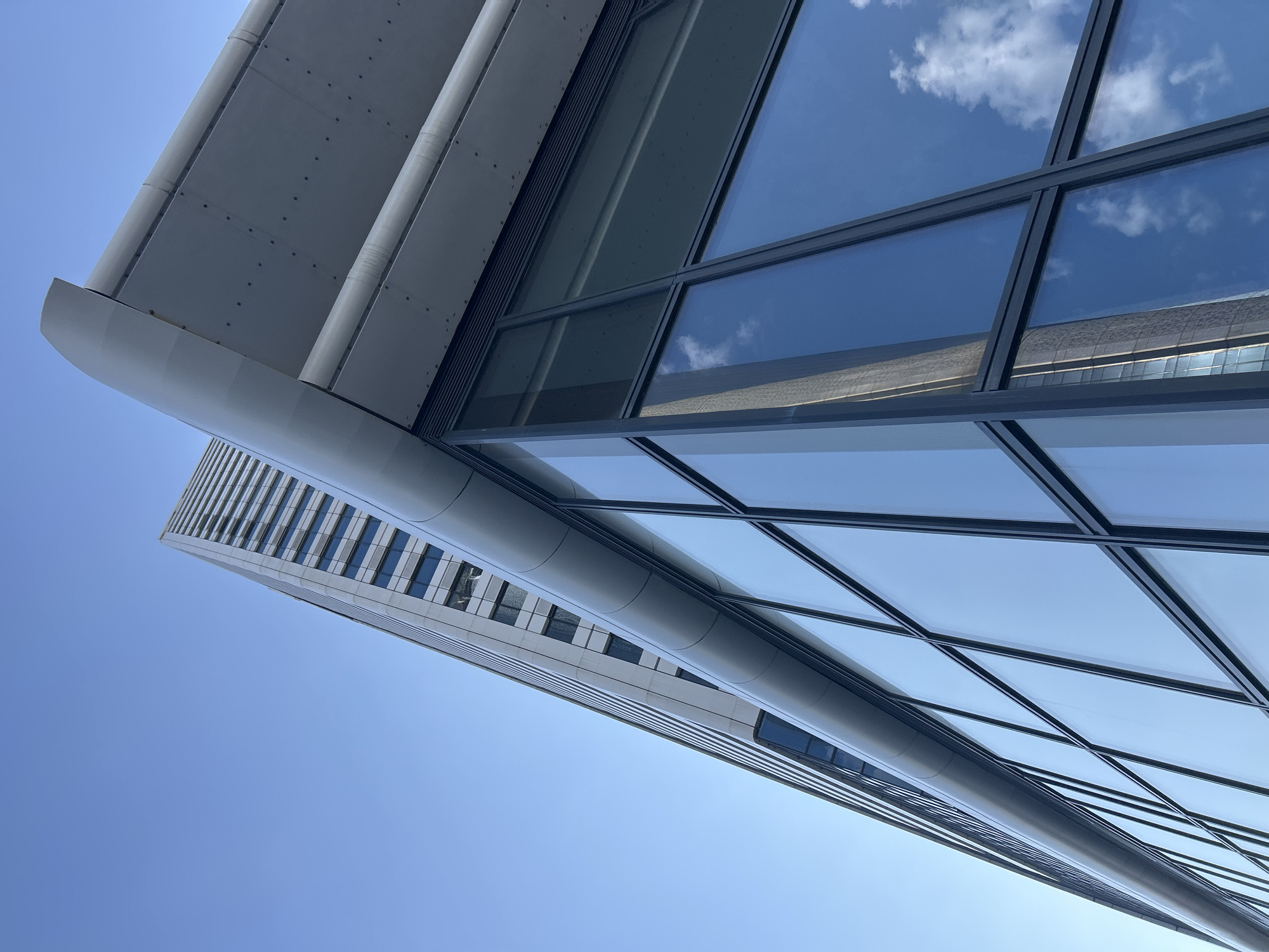 The image shows a low-angle, upward view of a modern skyscraper and an adjacent building with a glass facade, set against a clear blue sky. The skyscraper, in the background, features a regular pattern of windows. The foreground building has large, reflective glass panels that mirror the sky, clouds, and a portion of the skyscraper. The architectural style is contemporary, with clean lines and geometric shapes. The setting is an urban environment, likely in the city of Sofia, Bulgaria, given the context provided. There are no people visible in the image, and the scene is static and devoid of any action. The lighting suggests it is daytime, likely with bright sunlight.