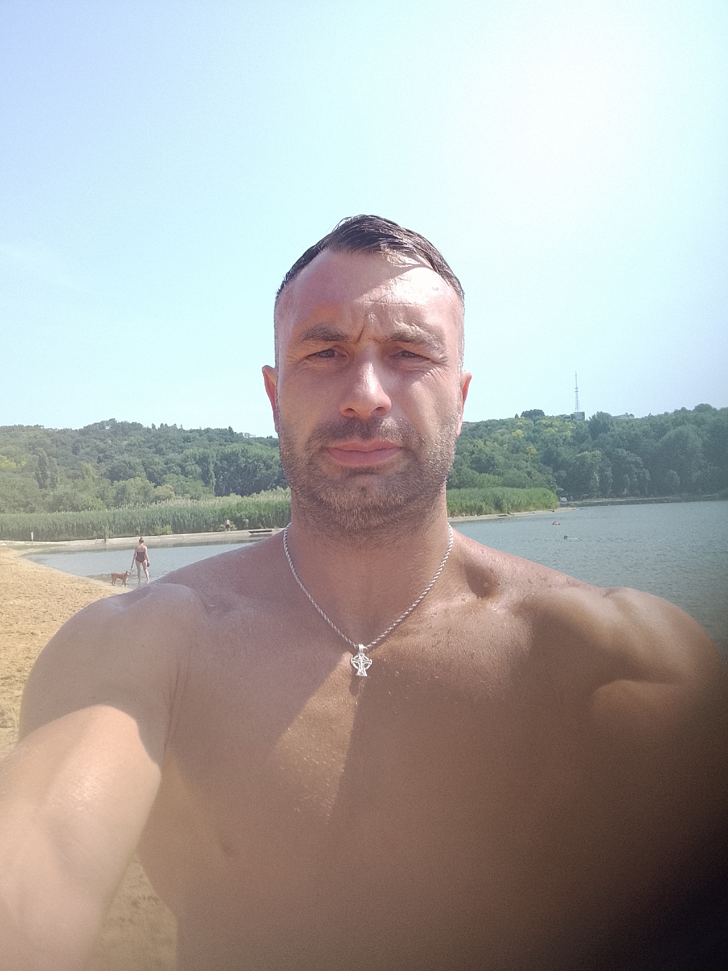 A man with a shaved head and stubble, wearing a silver chain with a Celtic knot pendant, takes a selfie on a sunny day at a lake. He is shirtless, and his skin appears wet. In the background, a sandy beach leads to a body of water with reeds along its edge. Across the water, a tree-covered hill rises, and a radio tower is visible on the summit. A woman with a dog is walking on the beach near the water's edge. The man's left arm is visible in the foreground, and the image is framed from the chest up. The location is not specified in the image.