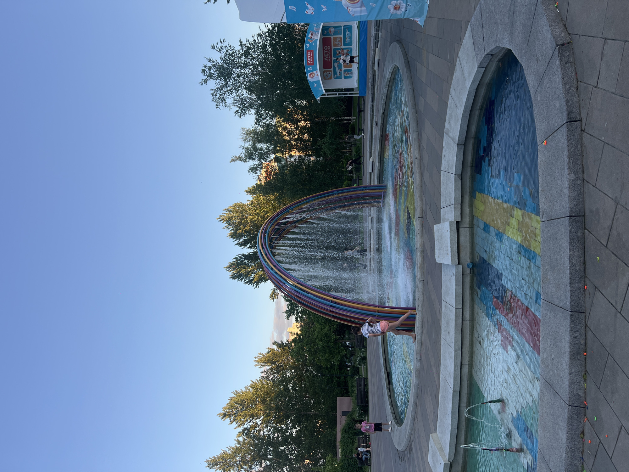 The image captures a scene in Moscow, Russia, featuring a colorful fountain with a rainbow-shaped arch. Water is flowing from the arch into a shallow pool adorned with mosaic tiles. A person stands at the edge of the fountain, interacting with the water. In the background, trees provide a green backdrop to the blue sky. A booth or stage with Russian text is visible near the fountain, and people are scattered nearby, some sitting on benches. The setting appears to be a public park or recreational area in Moscow.