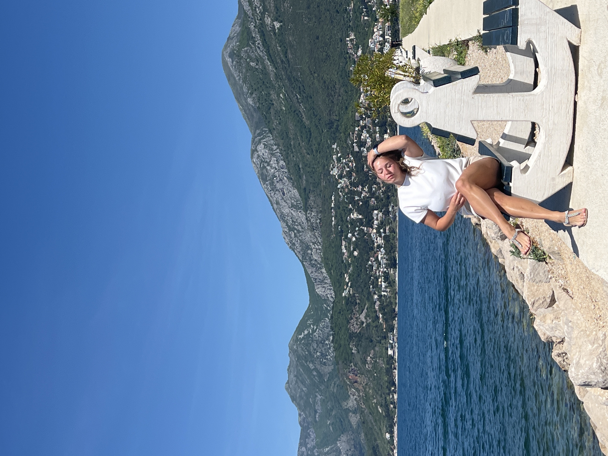 The image, taken on a bright, sunny day, captures a woman posing on a large, decorative anchor sculpture by the sea. The photo, currently rotated, would show a horizontal composition if corrected.
The main subject, a woman with light brown hair, is positioned on her side on the anchor. She is wearing a plain white t-shirt, light-colored shorts, and shiny silver sandals. A smartwatch is visible on her left wrist. Her eyes appear closed or squinting due to the intense sunlight, and one arm rests casually above her head, suggesting a relaxed posture.
The anchor, which doubles as a bench or seating area, is a prominent object in the foreground. It is crafted from a light-colored material, possibly concrete or stone, with dark blue or black sections forming the seat and backrest elements. This maritime-themed sculpture adds character to the waterfront setting.
Behind the woman and the anchor, a vast expanse of deep blue water stretches out, showing visible ripples on its surface. Beyond the water, majestic mountains rise, covered in lush green vegetation and dotted with numerous white and light-colored houses, indicative of a coastal town or settlement nestled into the hillside. The sky above is a uniform, vibrant blue, clear of any clouds, confirming the bright and sunny weather. The immediate ground beneath the anchor consists of natural, light-colored rocks.
The scene depicts a serene and picturesque coastal environment, evoking a sense of calm and leisure, likely at a scenic viewpoint in Bar, Montenegro. No visible text is present in the image.