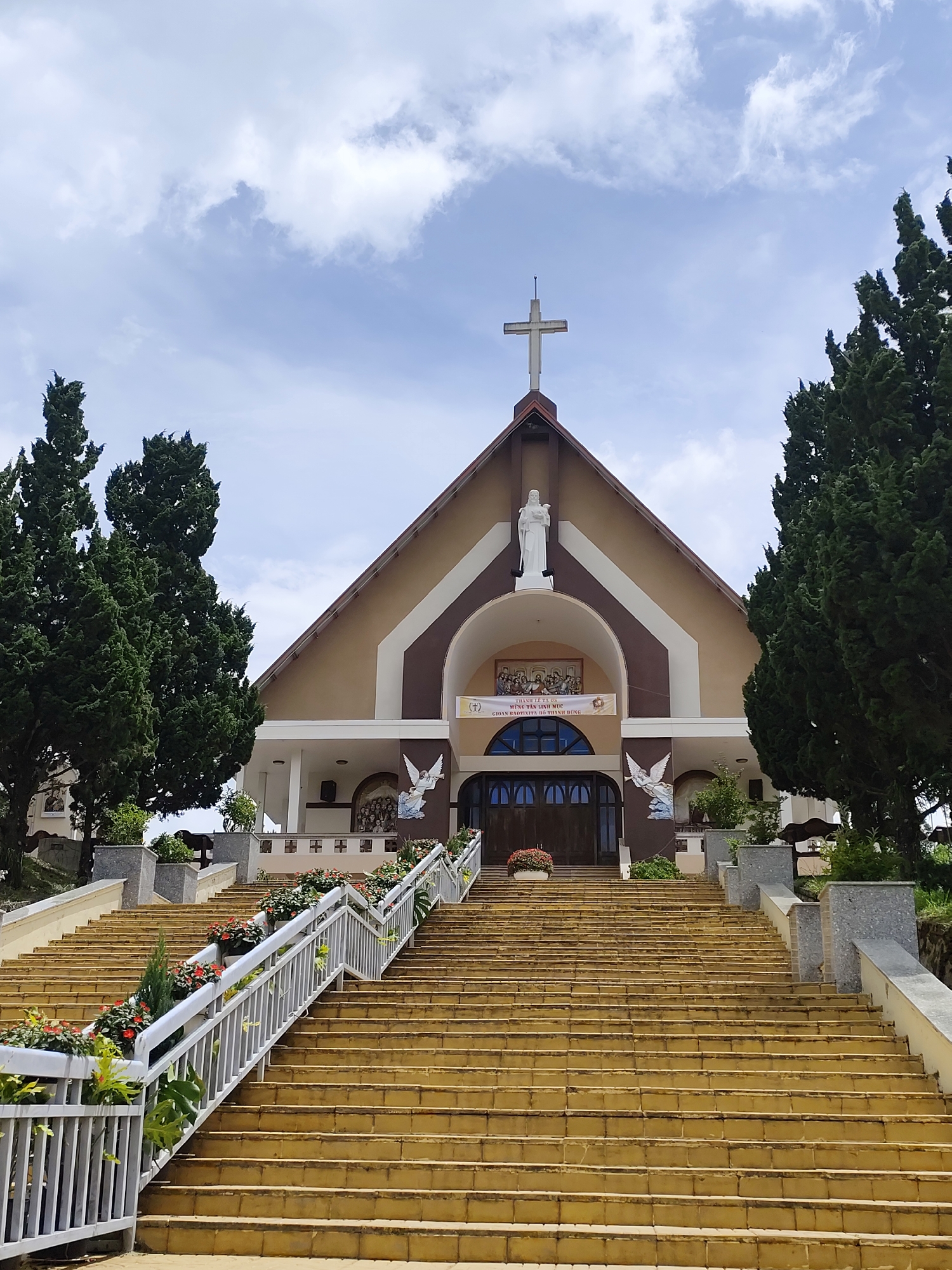 The image shows the entrance to a church, located in Lạc Dương, Vietnam. A wide, yellow staircase ascends to the church's main entrance. The staircase is flanked by white railings and planters filled with red and green foliage. At the top of the stairs, the church facade features a large arched doorway, two arched windows, and a white statue of a robed figure above the doorway. A large cross is mounted on the roof peak. Two statues of winged figures are visible on either side of the entrance. Tall green cypress trees stand on either side of the church, and the sky above is partly cloudy. A banner with Vietnamese text is displayed above the doorway.