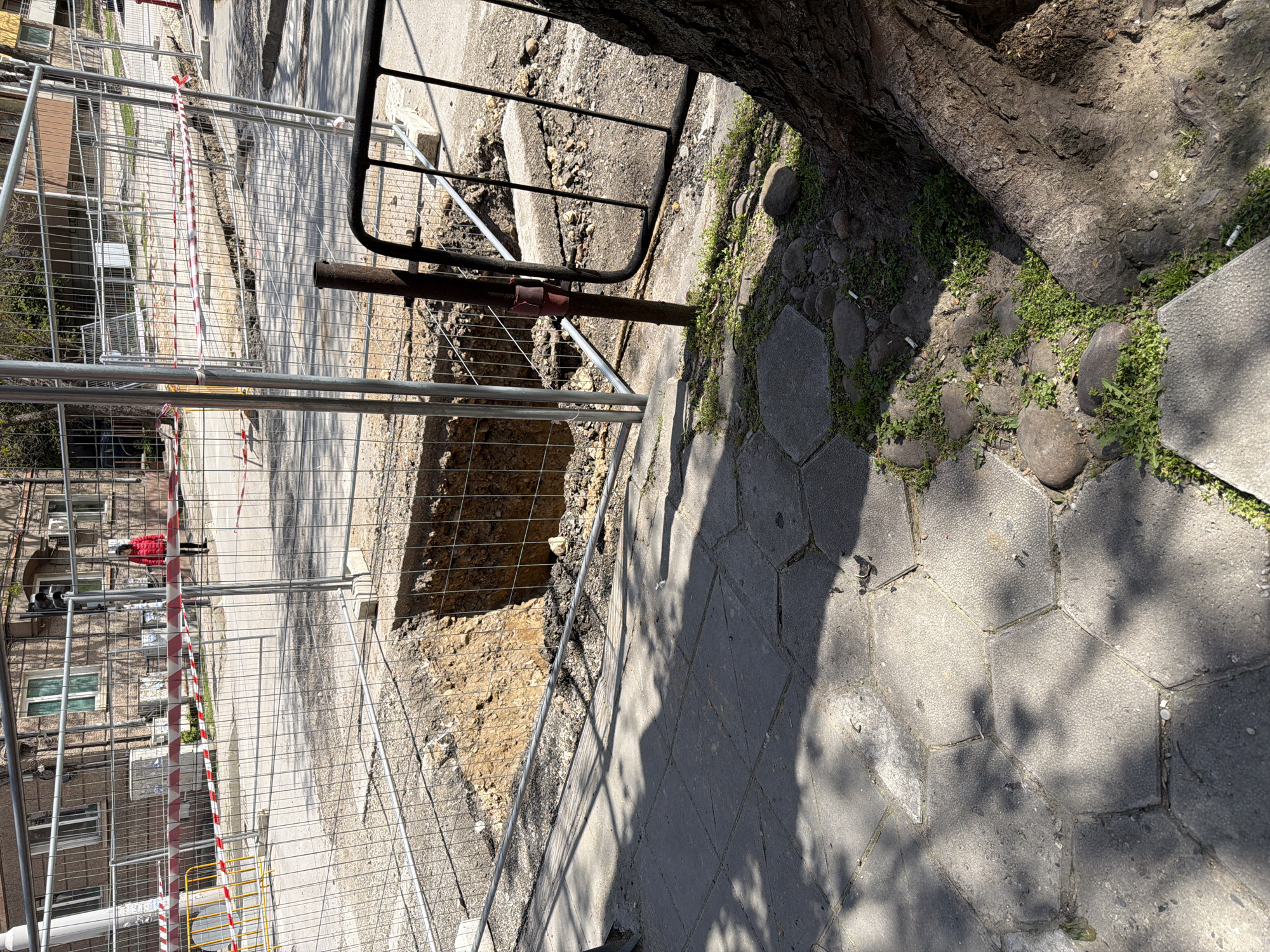 A construction site is located in Pleven, Bulgaria. A safety fence surrounds a trench on a paved sidewalk next to a tree. A person in a red jacket is visible in the background behind the fence.