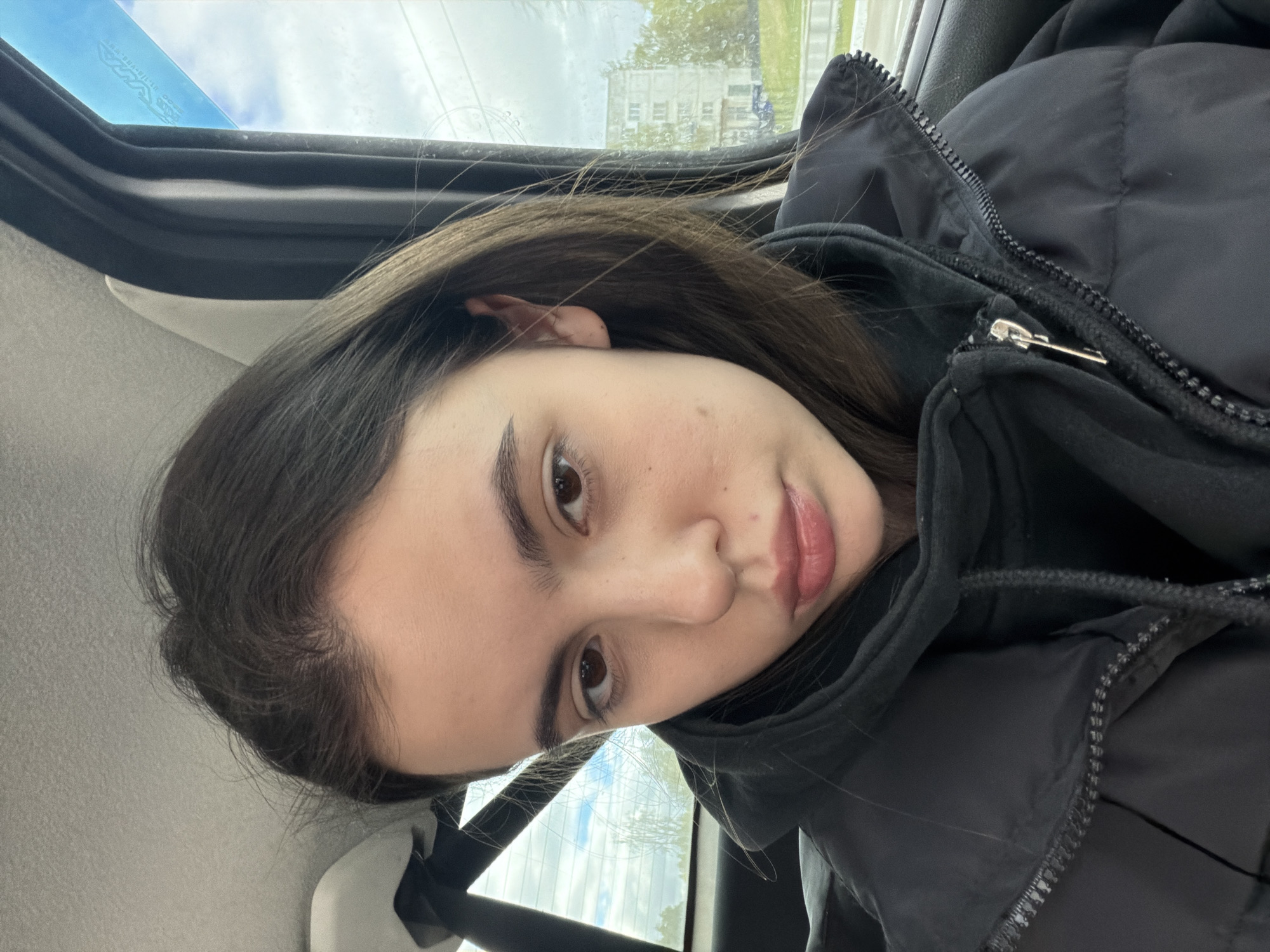 A young woman is seated in a vehicle, likely a car, as evidenced by the window frame and interior. She is wearing a black hooded jacket, and the zipper is partially visible. The background visible through the car window shows a building and vegetation. Given the context, the location is Chaikovsky, Russia. The image appears to be a casual selfie.
