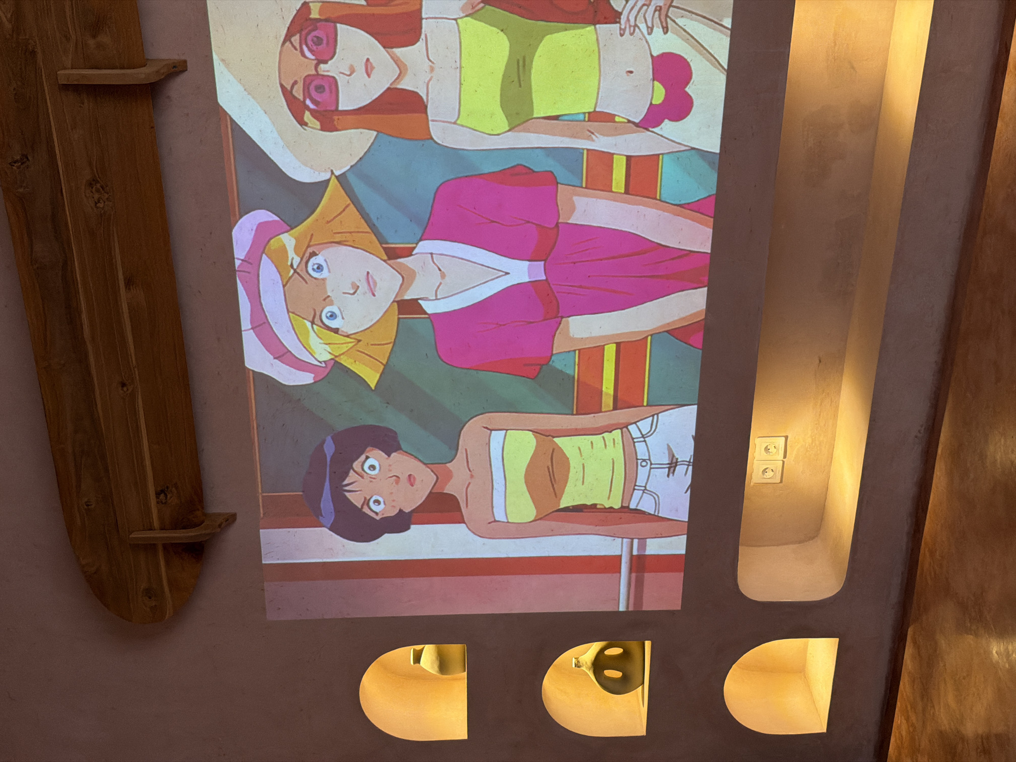 The image shows a wall in a building in North Kuta, Indonesia. The wall is painted a tan color and has a poster of the cartoon "Totally Spies" affixed to it. To the left of the poster is a wooden object, possibly a shelf. To the right of the poster is a recess in the wall where there are two electrical outlets. Below the poster are three smaller recesses in the wall with light shining from them.